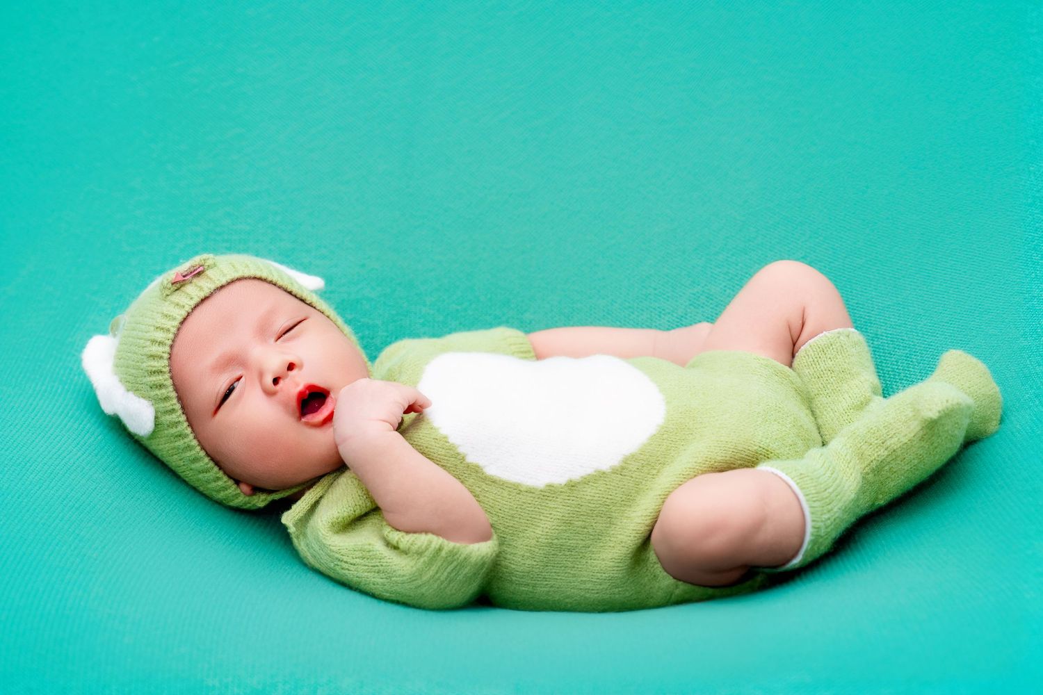 A newborn wearing a mint green costume lying on turquoise background.