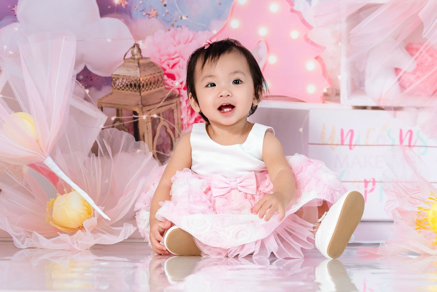 A toddler in white dress sits on pink floor surrounded by dreamy decorations.
