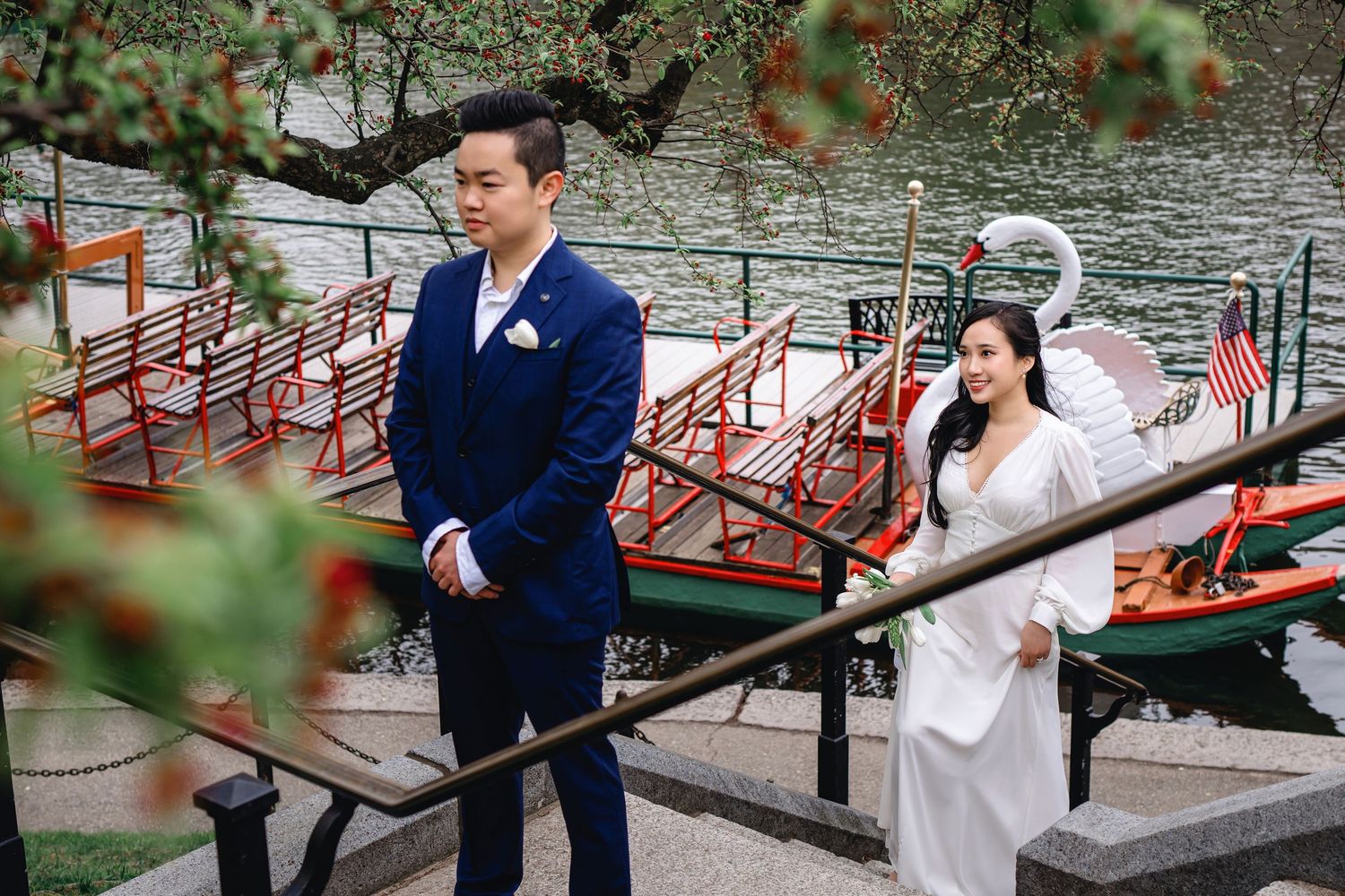 A couple in formal attire poses together on wooden docks near boats and red railings by a tranquil lake.