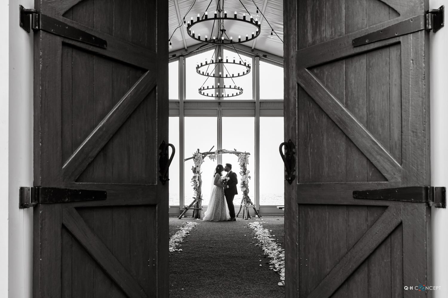 A dramatic black and white wedding photo framed through rustic barn doors with a chandelier overhead.