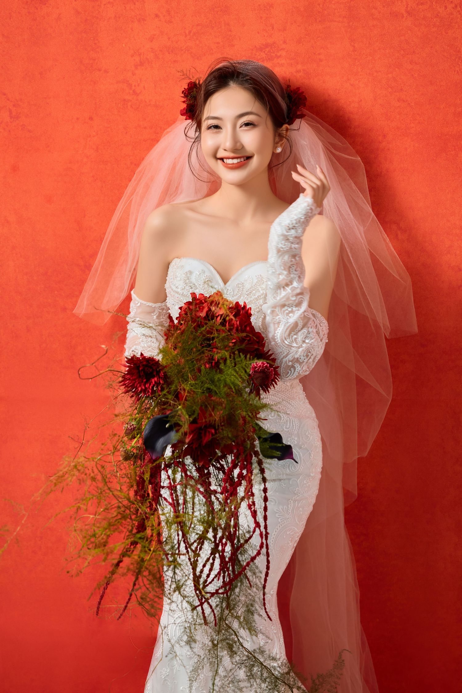 Bride in white lace dress poses with dramatic red floral bouquet against coral backdrop.