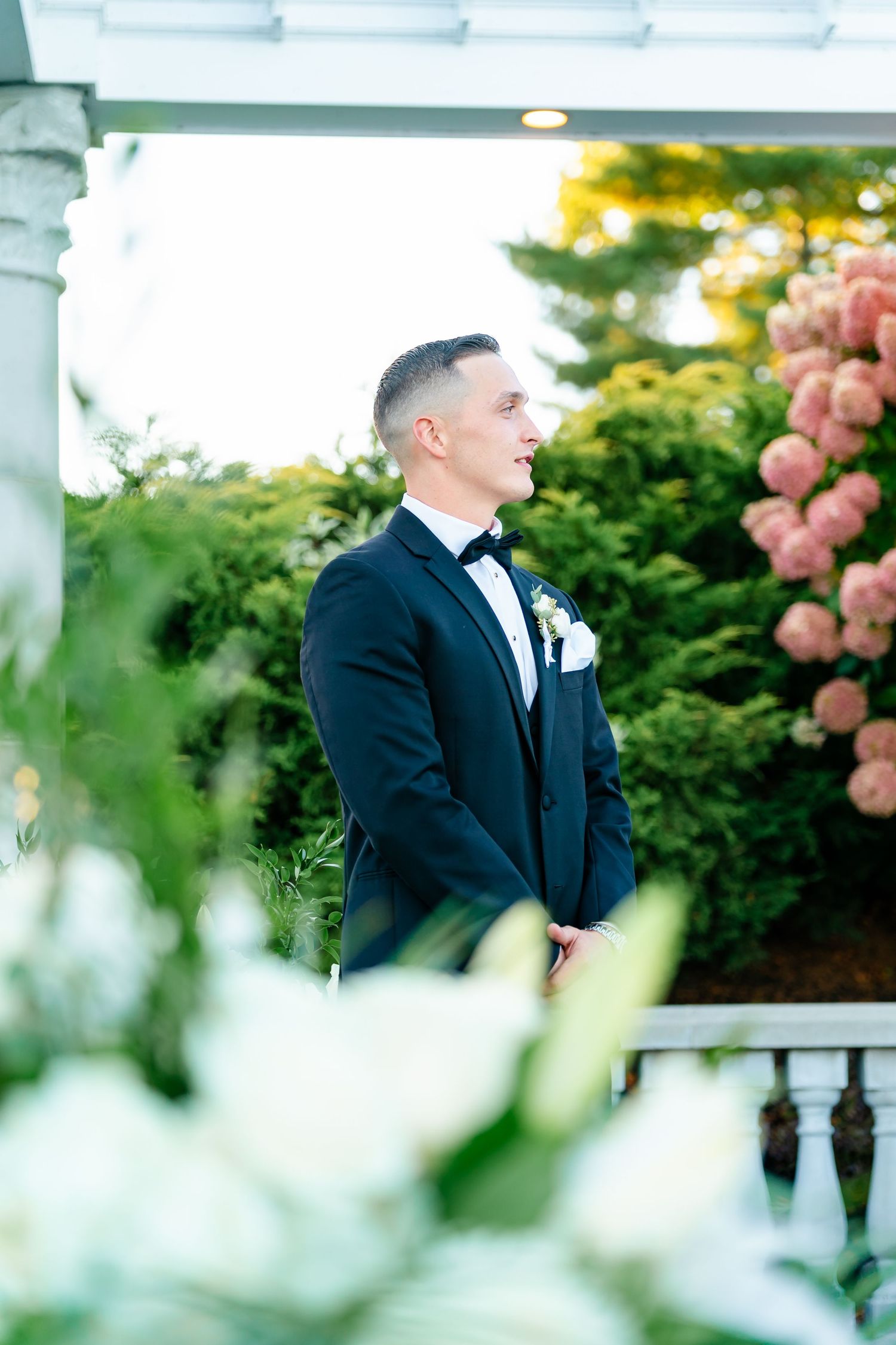 A groom in a classic black tuxedo smiles during an outdoor wedding ceremony with pink flowers in the background.