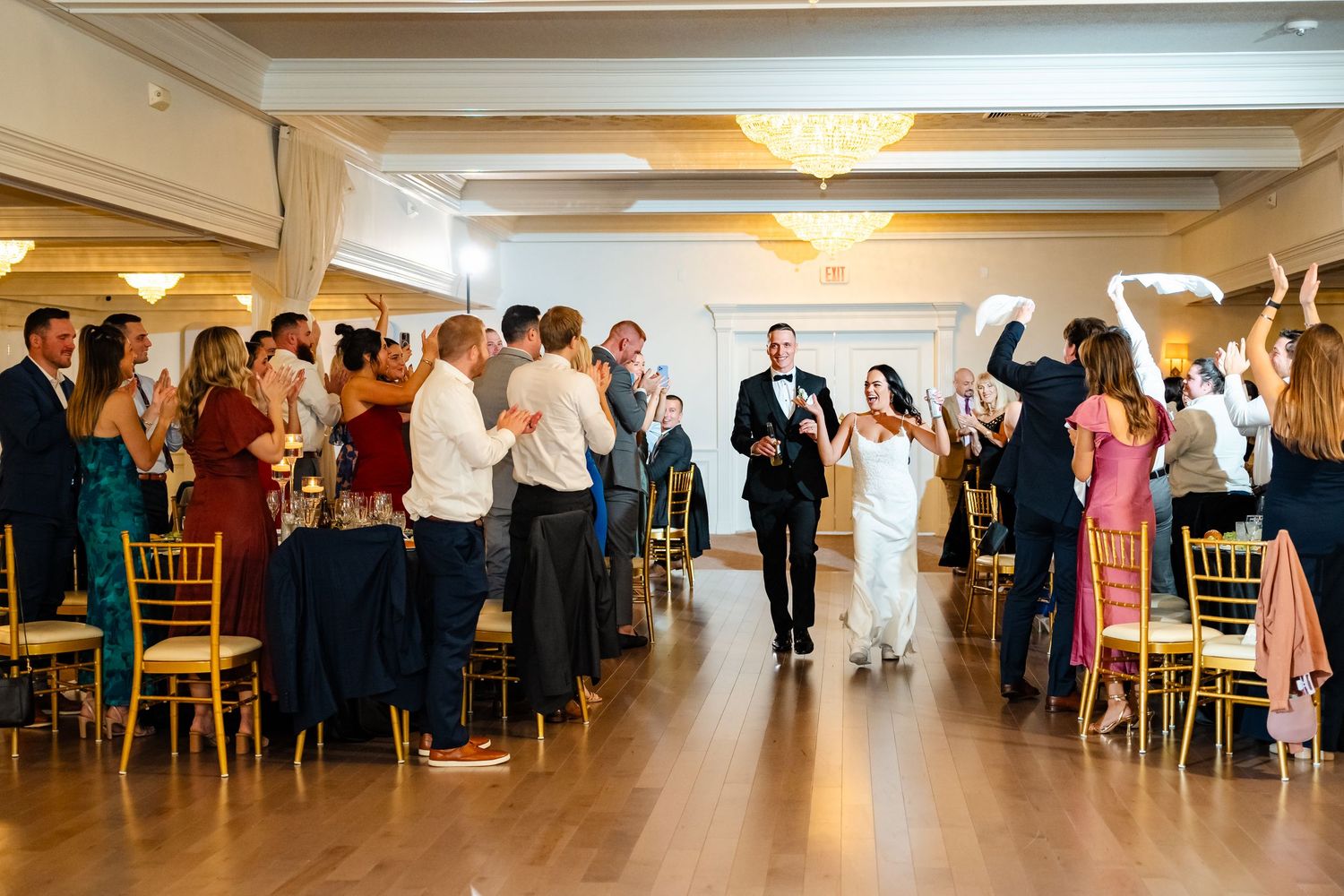 Wedding guests line up in an elegant reception hall with chandeliers and wooden chairs.