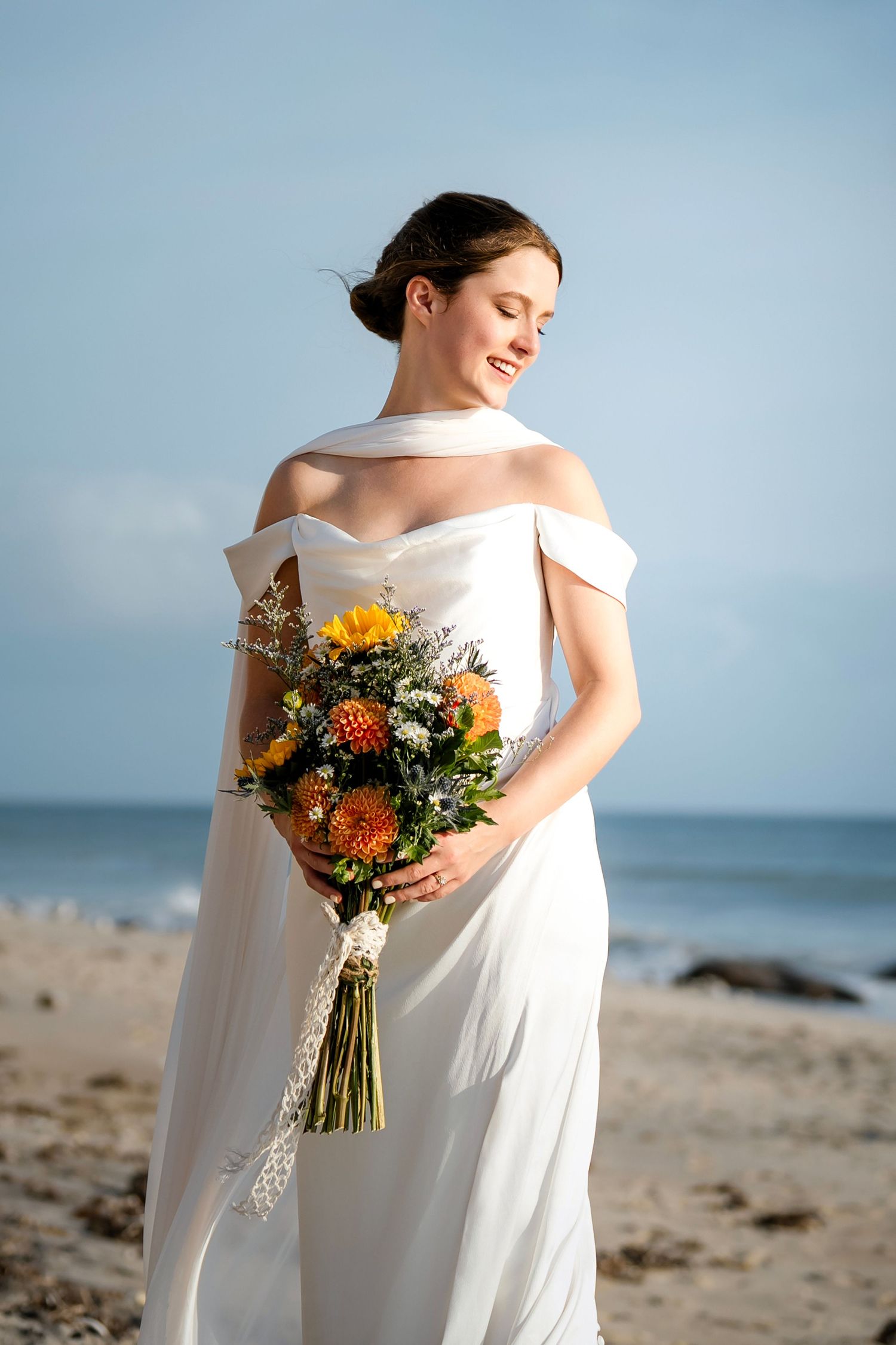 Bride in an elegant white off-shoulder dress holding a vibrant orange and yellow bouquet on the beach.