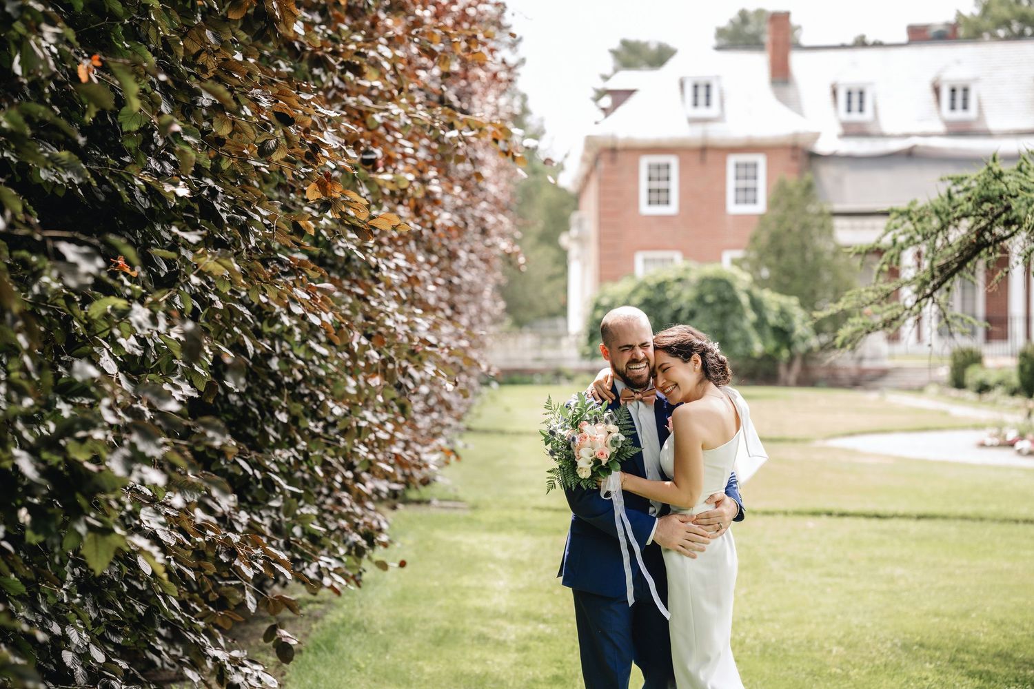 A couple shares a romantic moment on a sunny lawn surrounded by autumn trees and brick buildings.