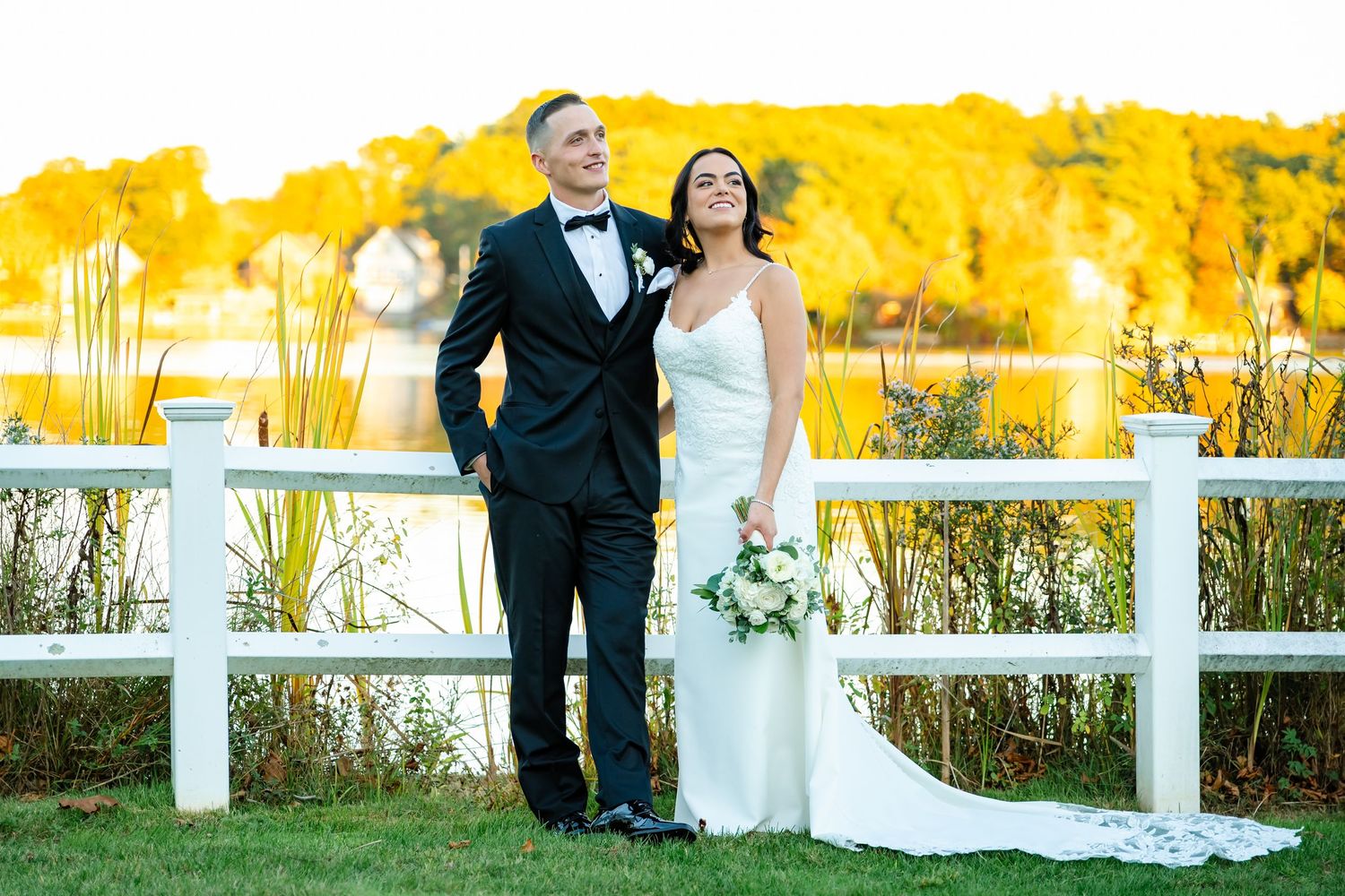 Couple poses by white fence with vibrant yellow autumn foliage in background during sunset.