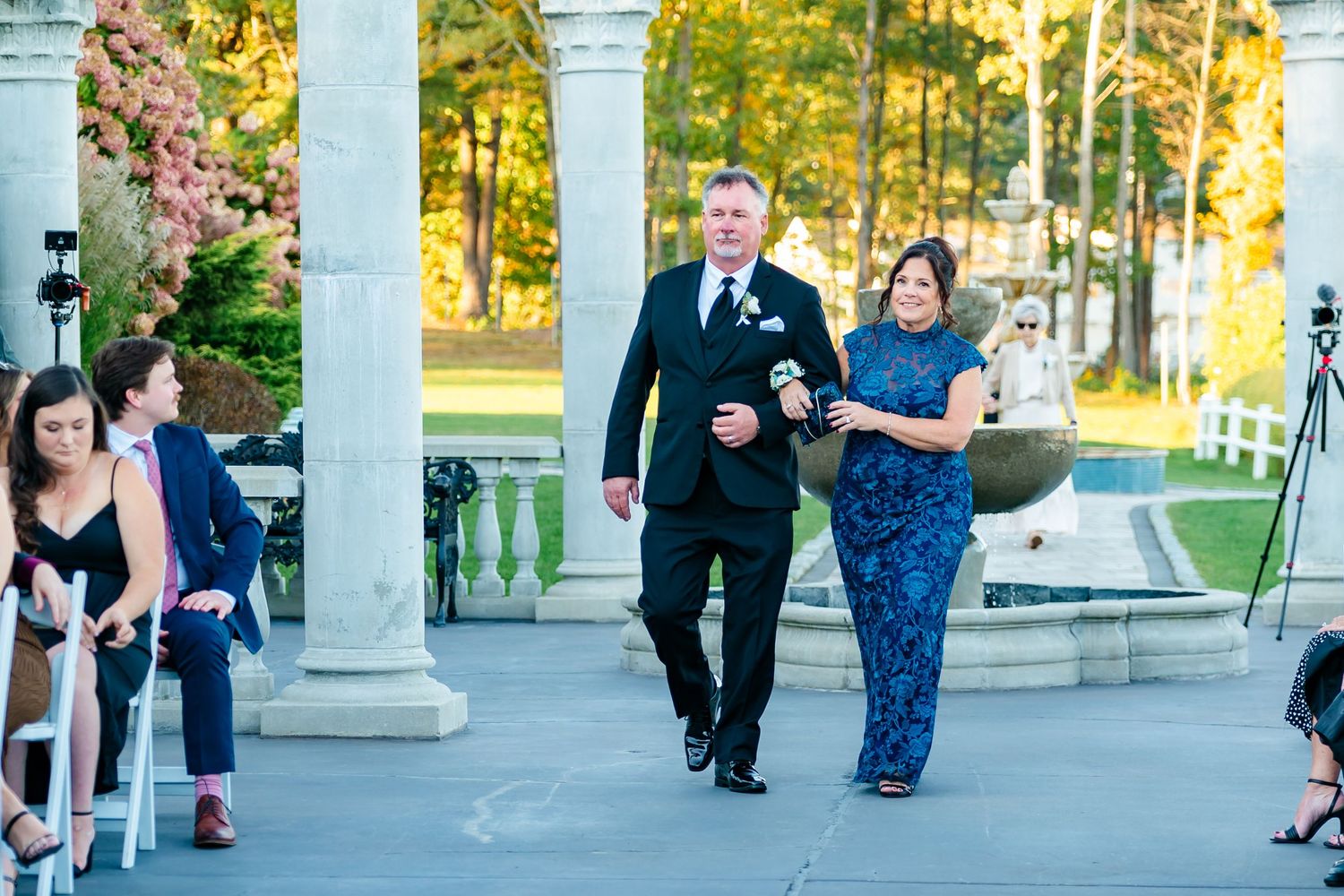 Outdoor wedding ceremony setting with white columns and guests gathering in late afternoon sunlight.