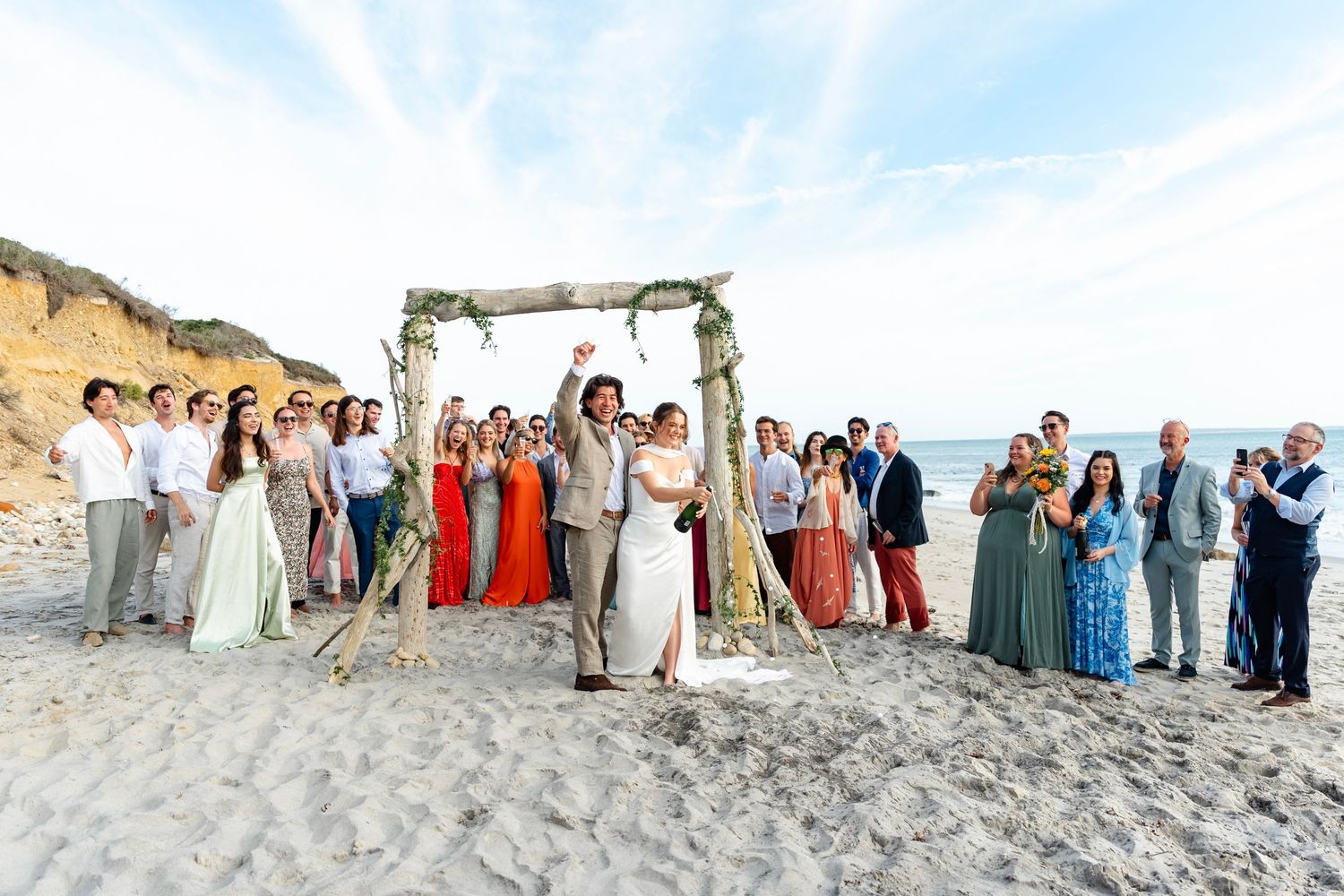 Wedding guests lined up on a sandy beach with cliffs and ocean in the background.