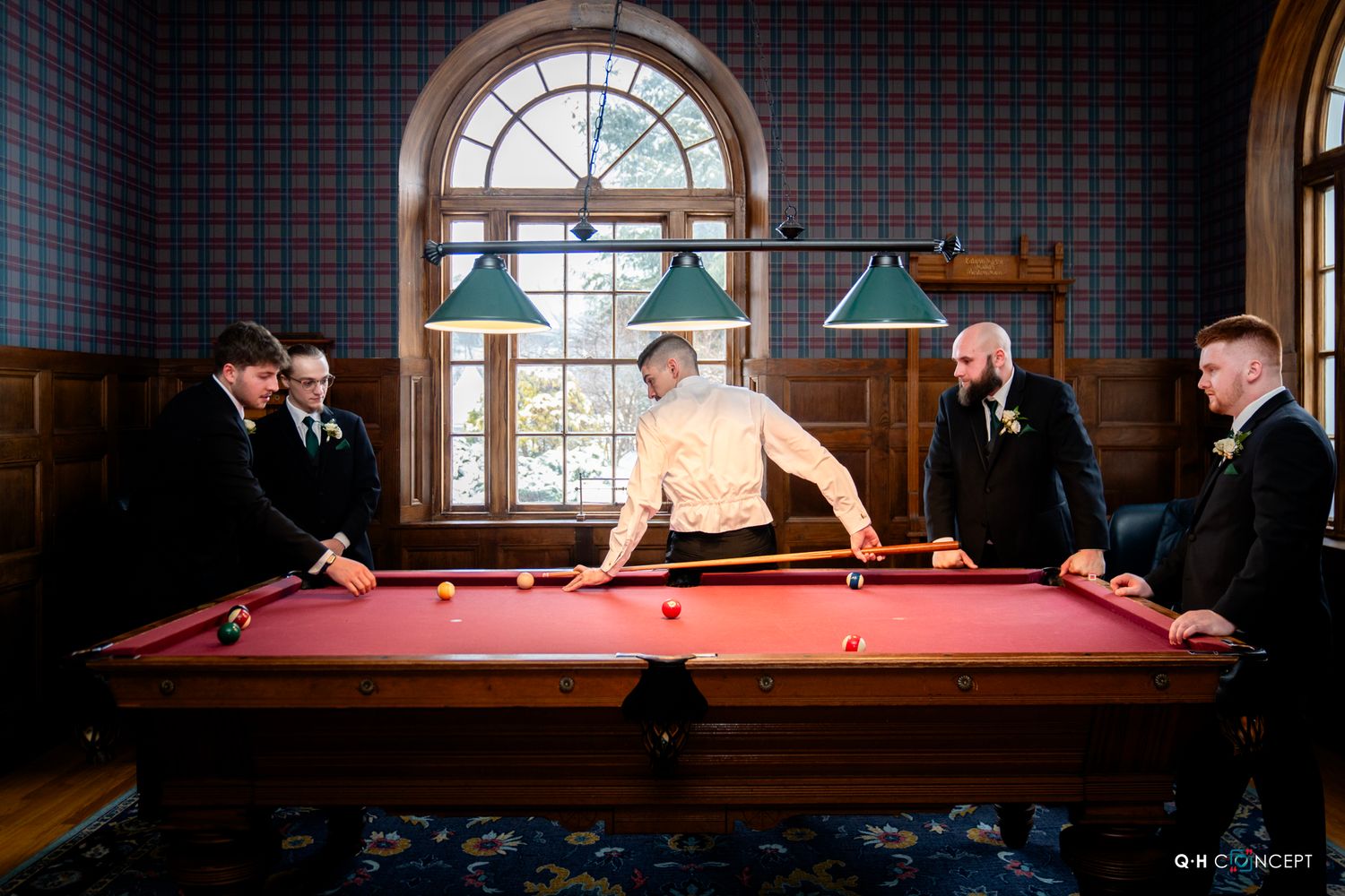 Groomsmen gather around a pool table in a wood-paneled room with an arched window.