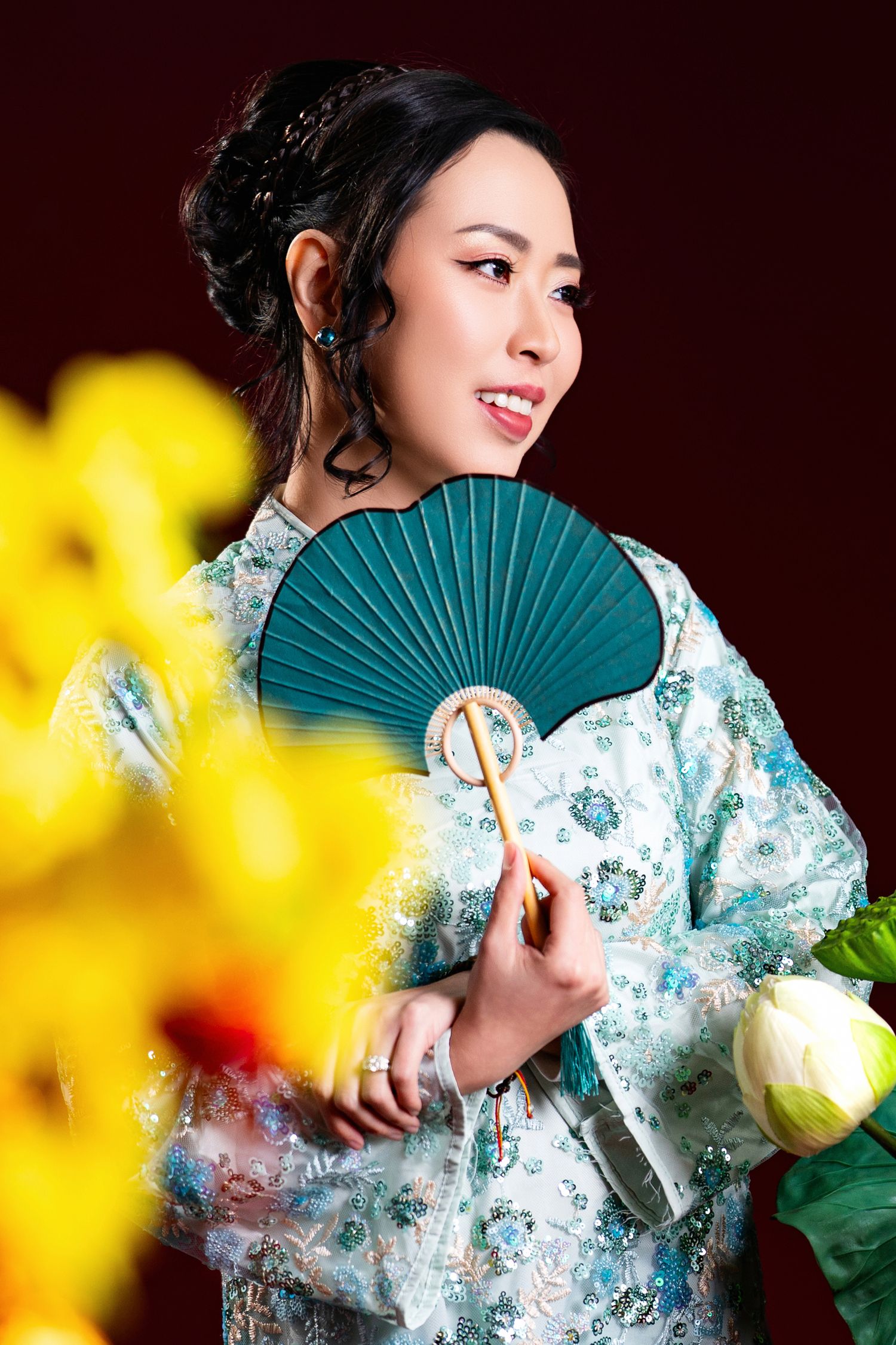 Traditional Asian attire with teal fan poses against dark background with yellow flowers in foreground.