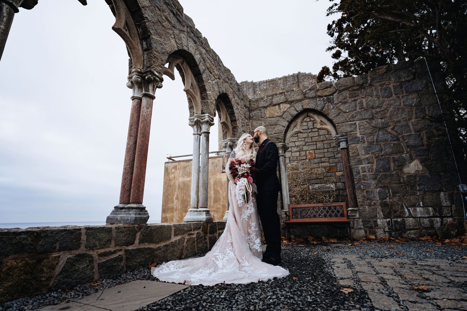 A couple embraces under stone archways at a historic venue with dramatic cloudy skies in the background.