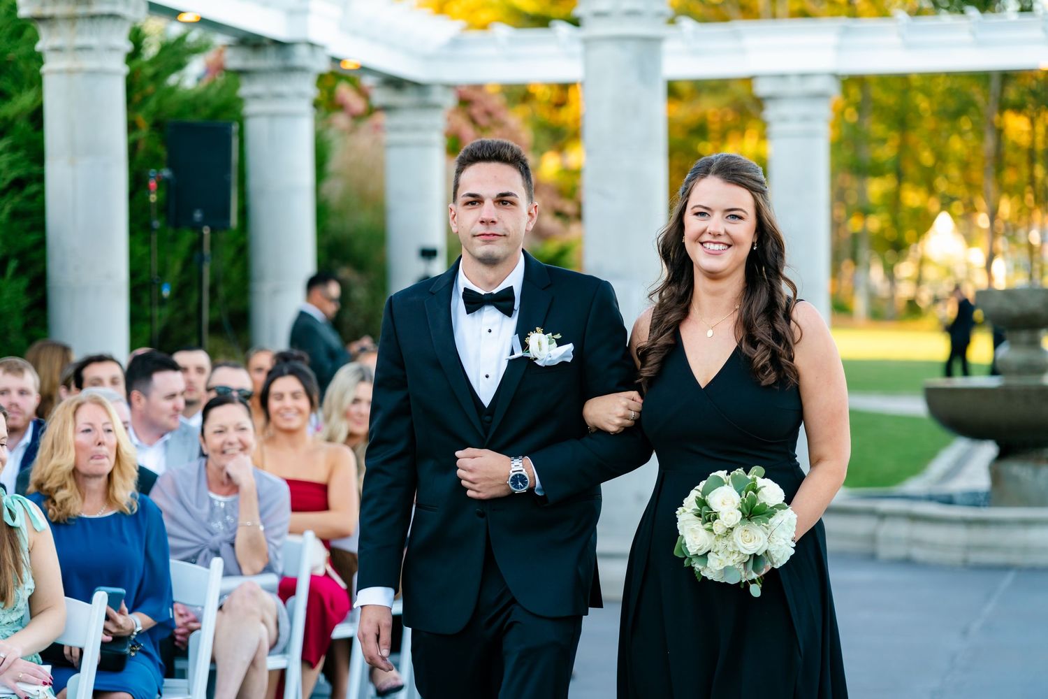 Wedding party member escorts bridesmaid down aisle during outdoor ceremony with fountain backdrop.