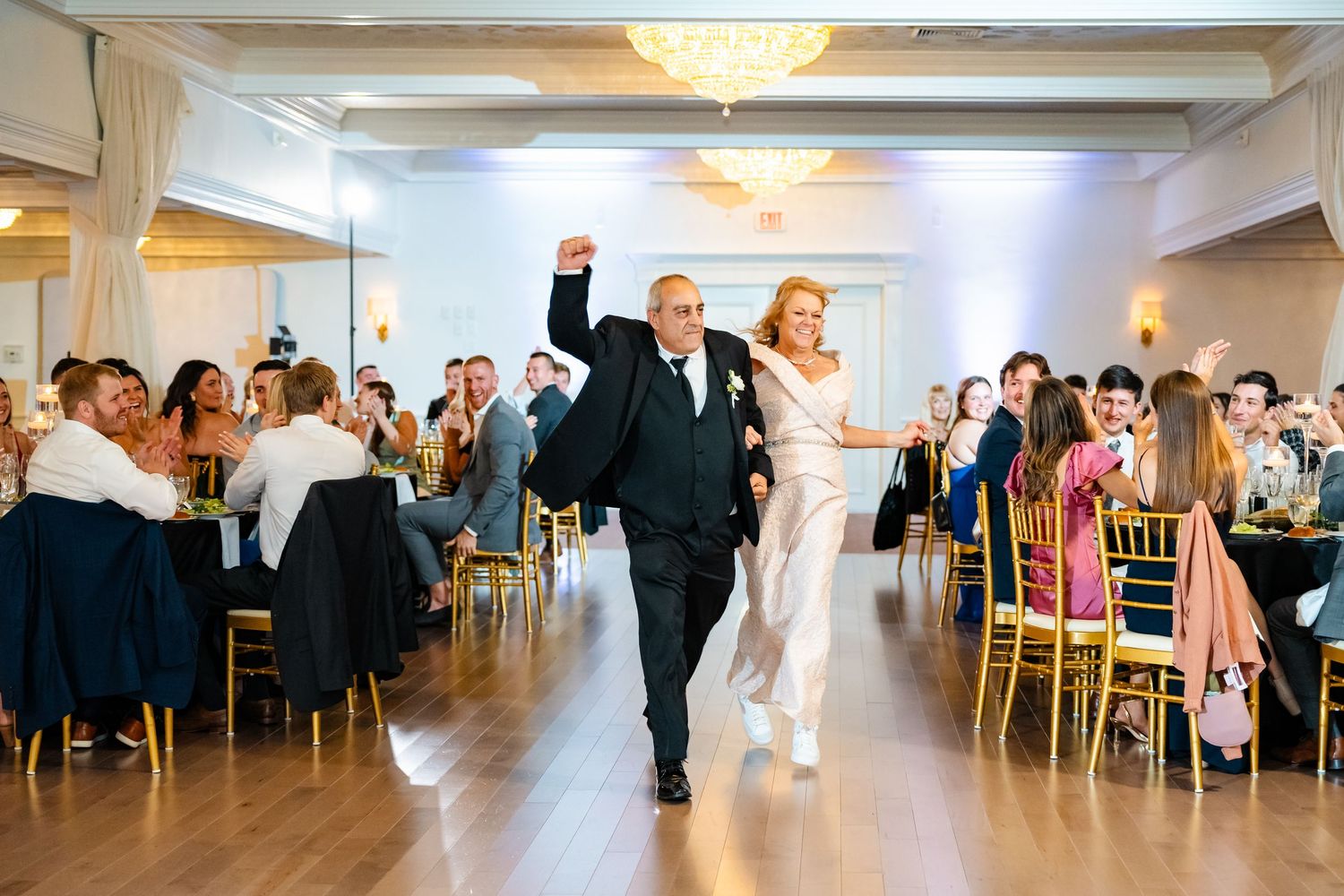 Wedding guests dancing and celebrating in an elegant ballroom with chandeliers and white decor.