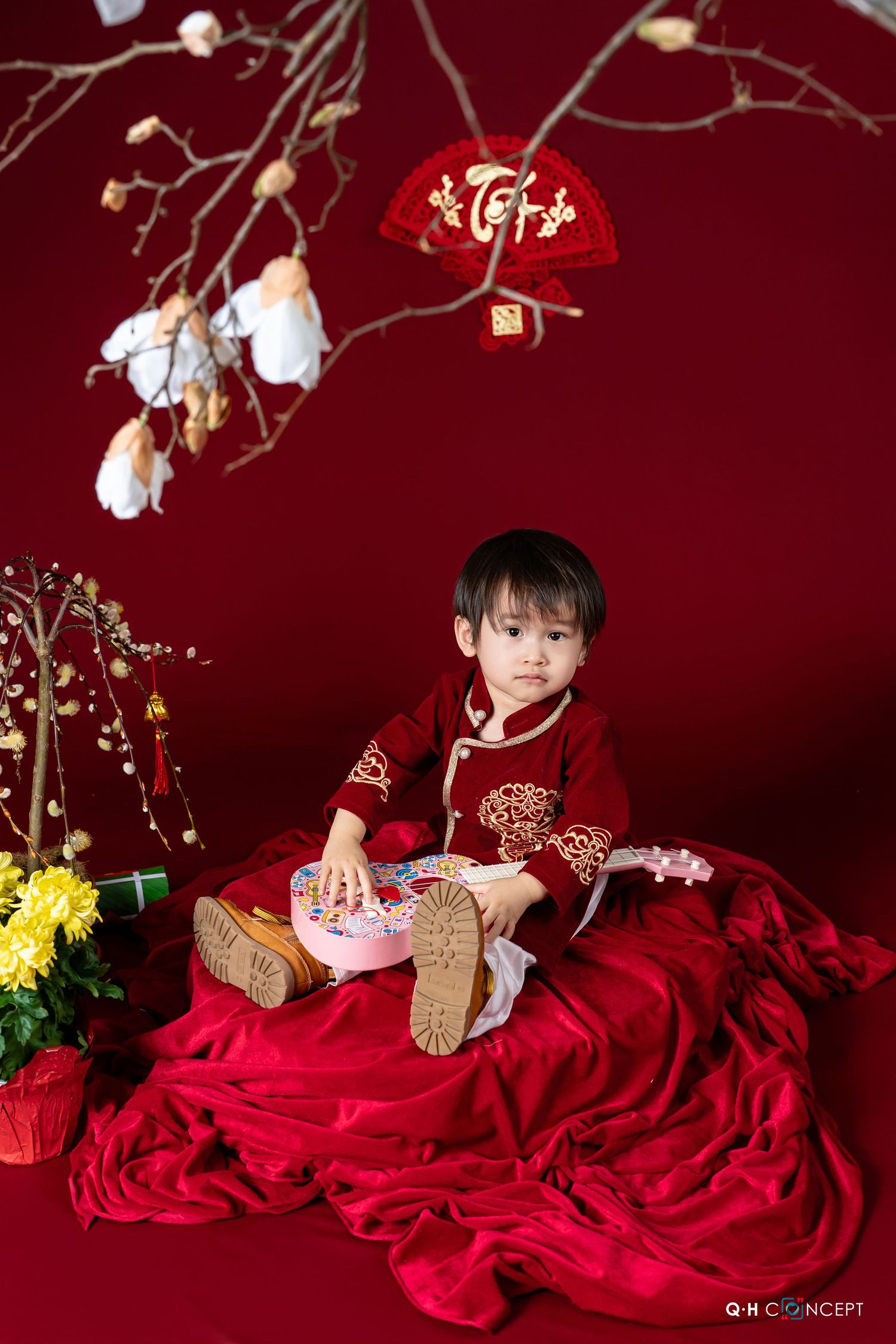 A child in a flowing red dress lies on a dark backdrop surrounded by delicate white floral decorations and branches.