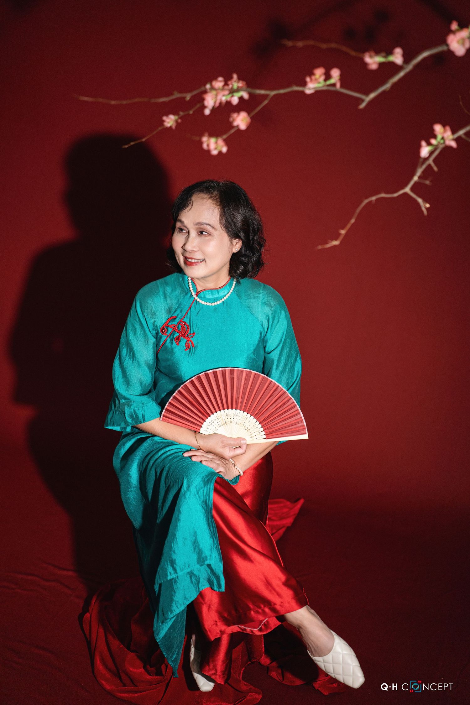 Person in turquoise traditional Asian dress holding red fan poses against red backdrop with cherry blossom branch.