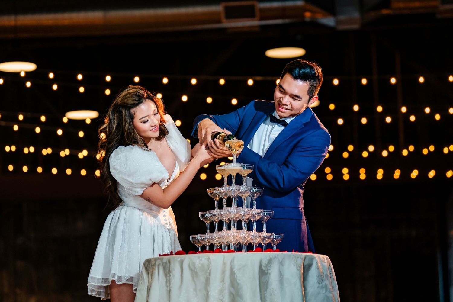 Couple shares playful moment near wedding cake with string lights in background.