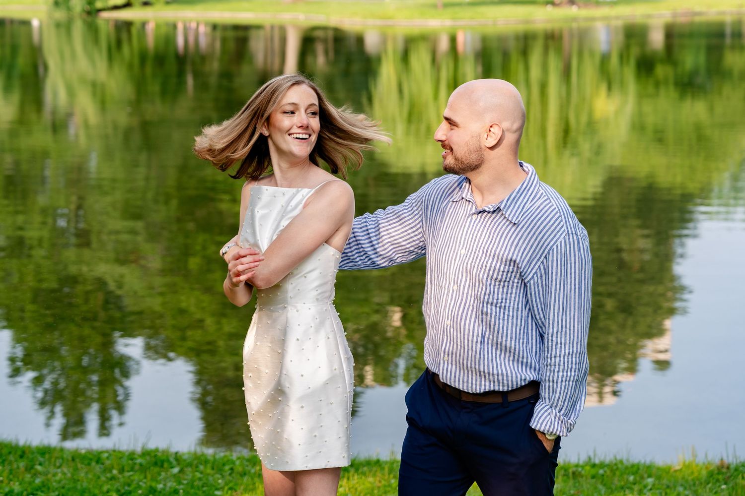 A smiling couple dancing by a serene lake, woman in white dress with flowing hair, man in blue striped shirt.