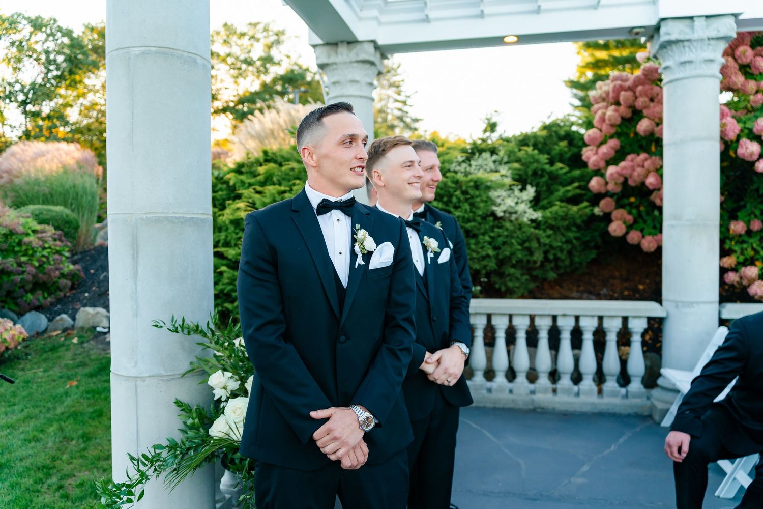 Two people in matching black tuxedos stand together under a white pergola during an outdoor ceremony.