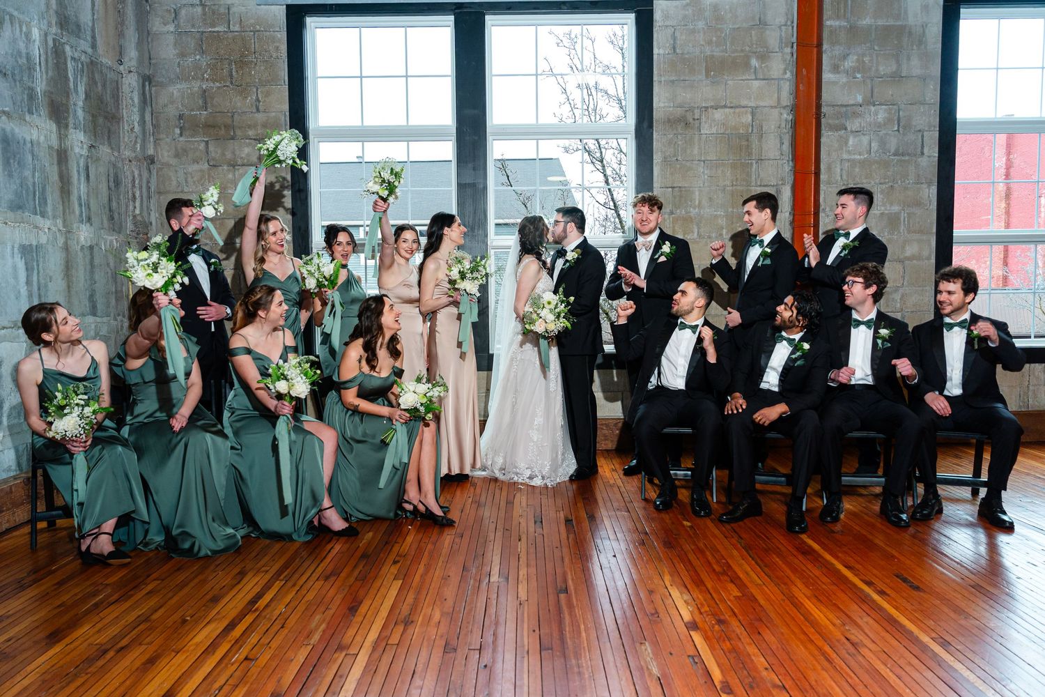Wedding party lined up against brick wall windows in formal attire.