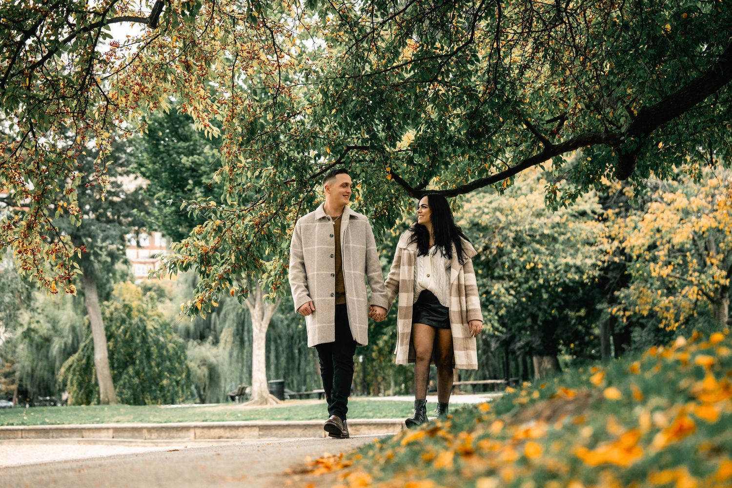 A couple walks together through a scenic park path lined with blooming yellow flowers and lush green trees.