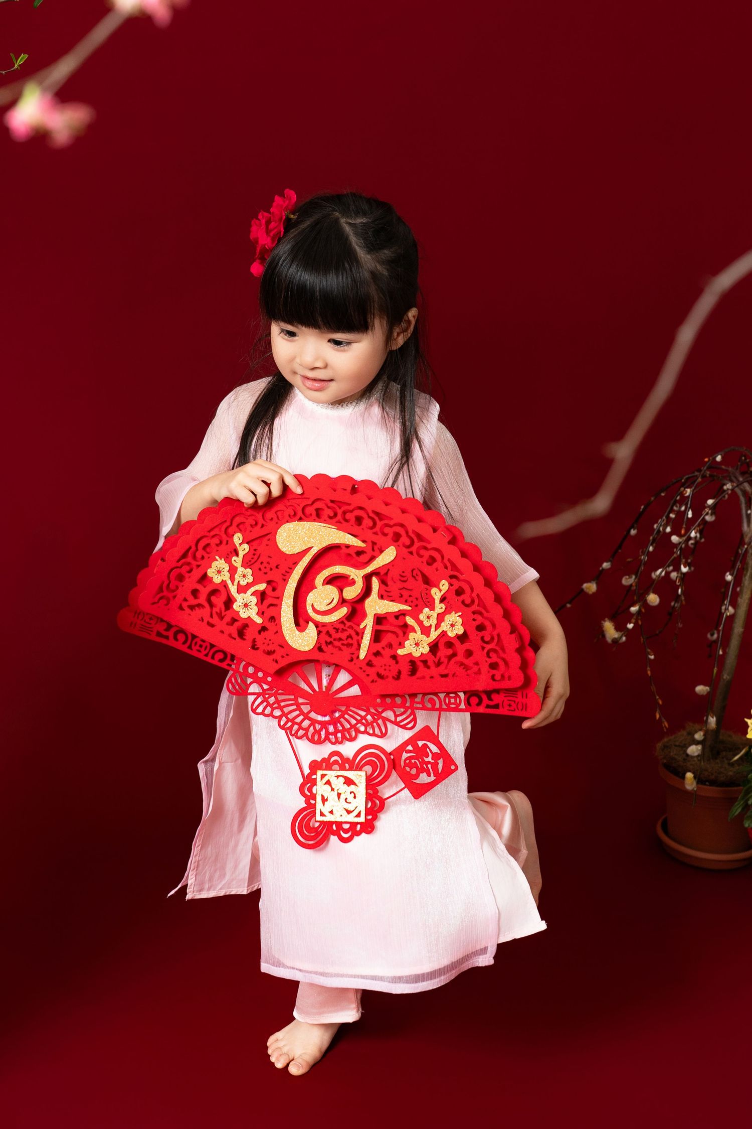 A young child in a white dress holds a decorative red Chinese fan against a deep red background with floral accents.