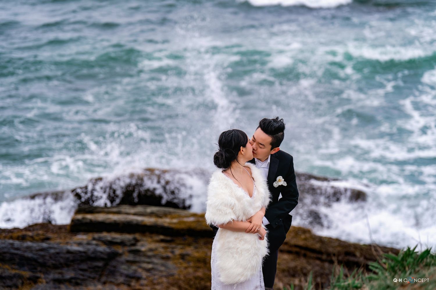 A couple in wedding attire embrace on rocky coastal cliffs with dramatic crashing waves in the background.