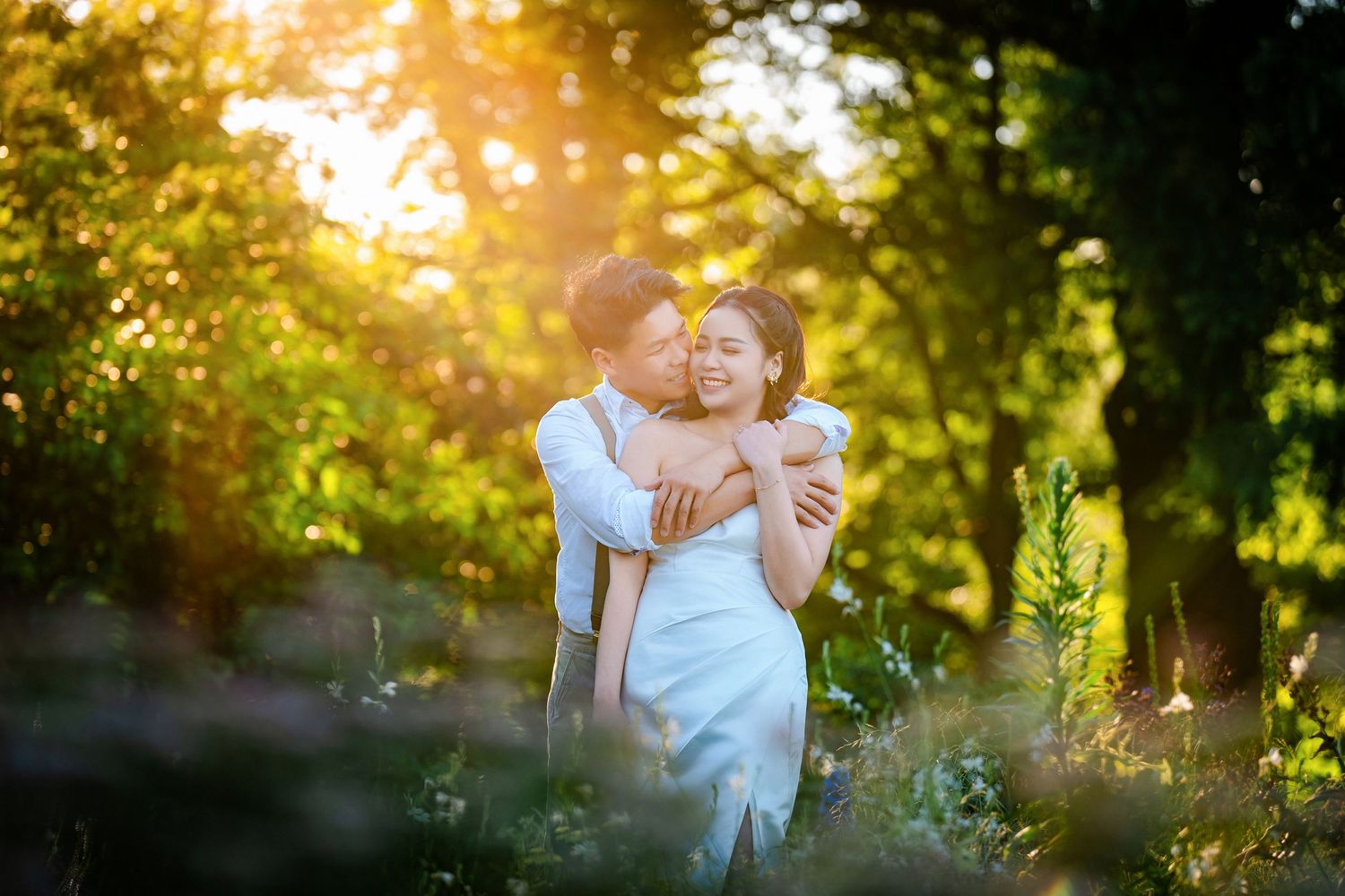 Couple embracing in a sunlit garden with golden hour light filtering through the trees.