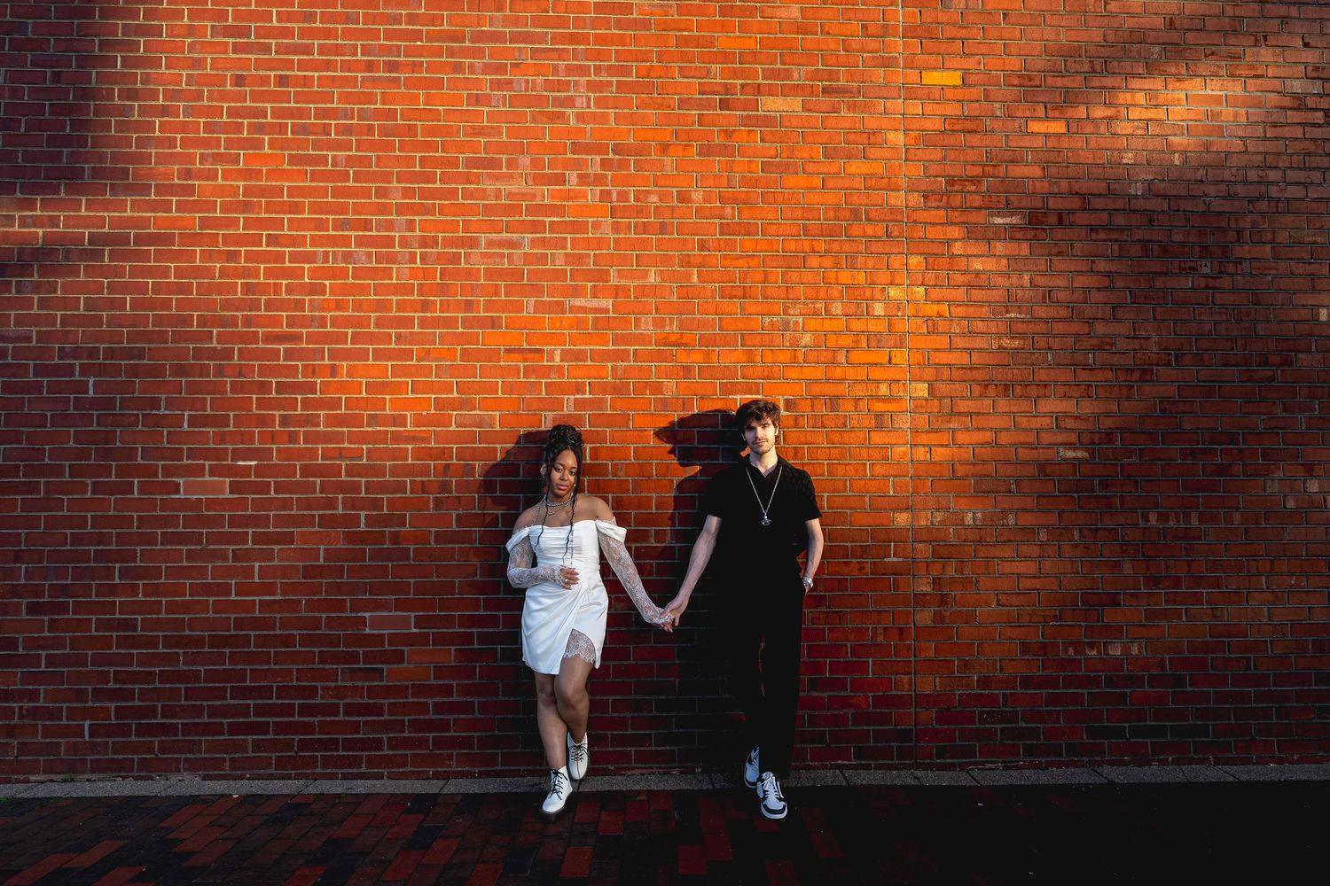 Two people holding hands and leaning against a red brick wall, one wearing white and one wearing black.