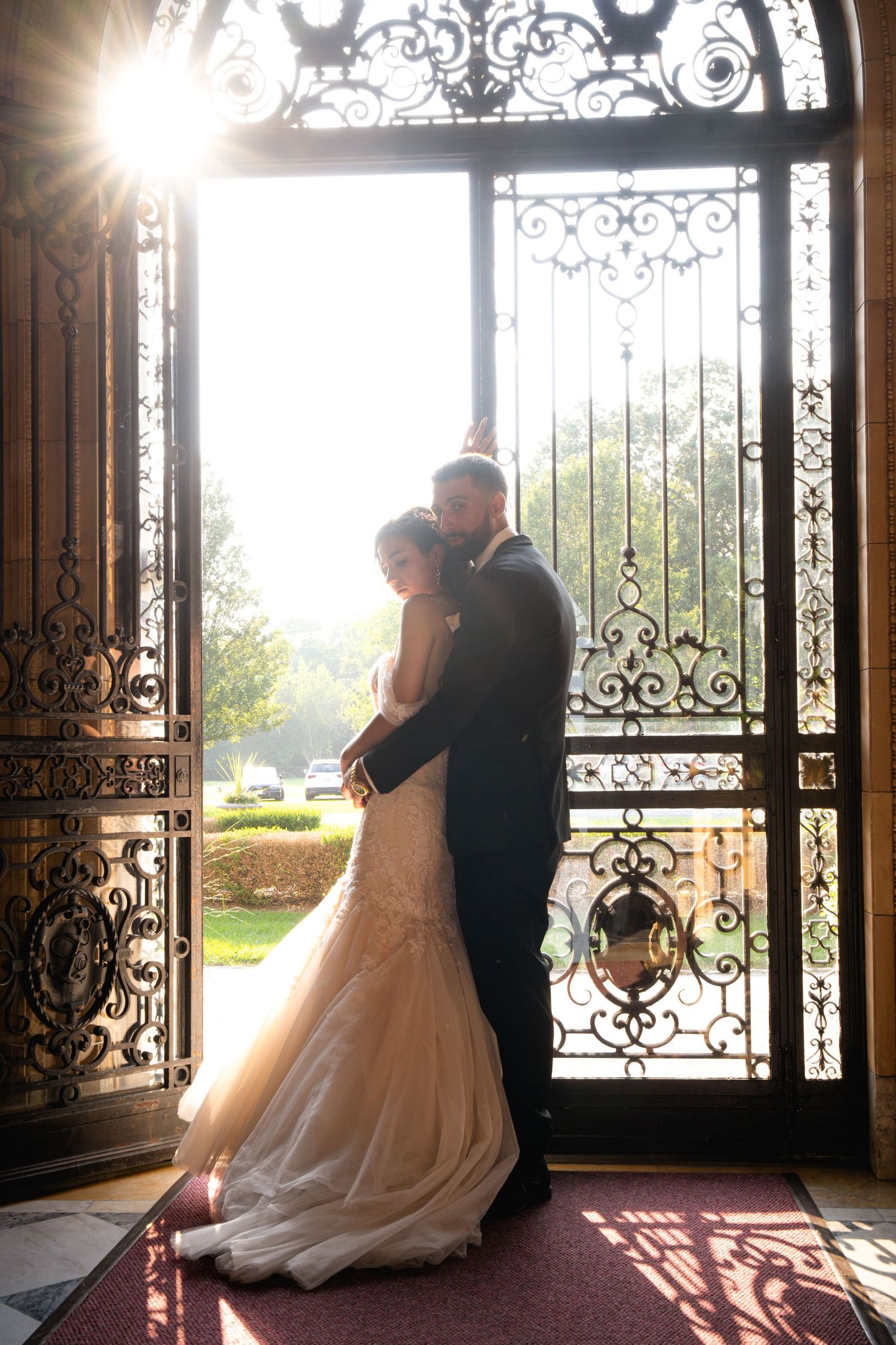 Silhouetted couple shares romantic moments against ornate iron doors with natural sunlight streaming through windows.