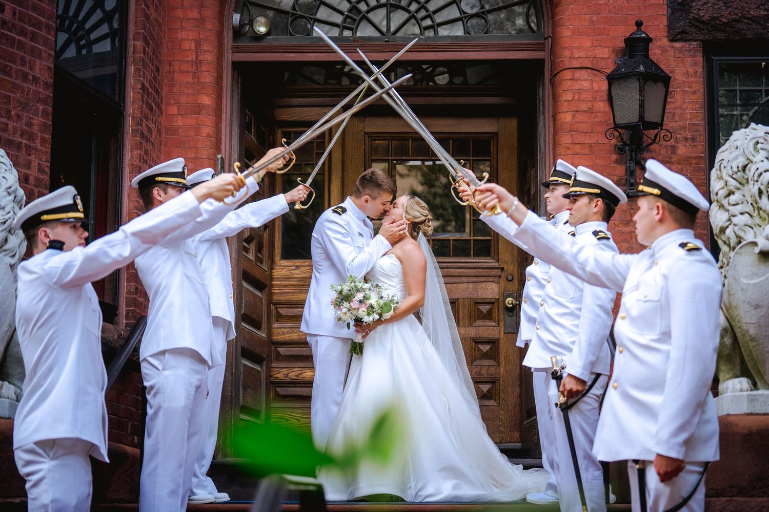 Wedding party members in white uniforms perform a synchronized dabbing dance move in front of a brick building.