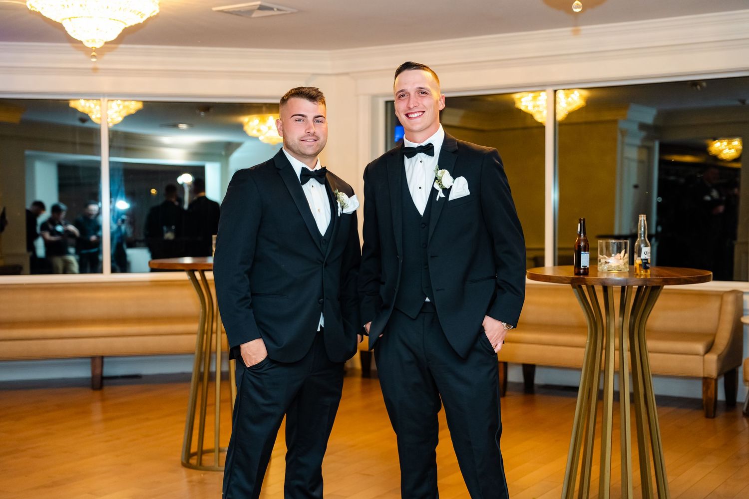 Two people in black tuxedos pose in elegant ballroom setting.