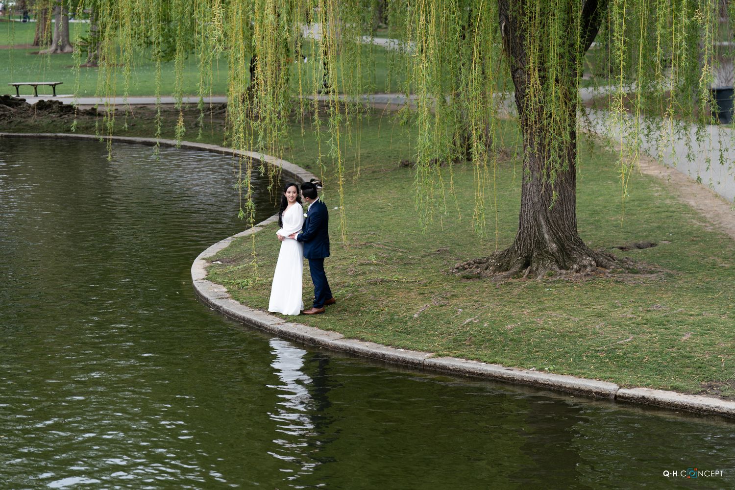 Bride and groom pose romantically on a curved stone path by a tranquil pond surrounded by weeping willows.
