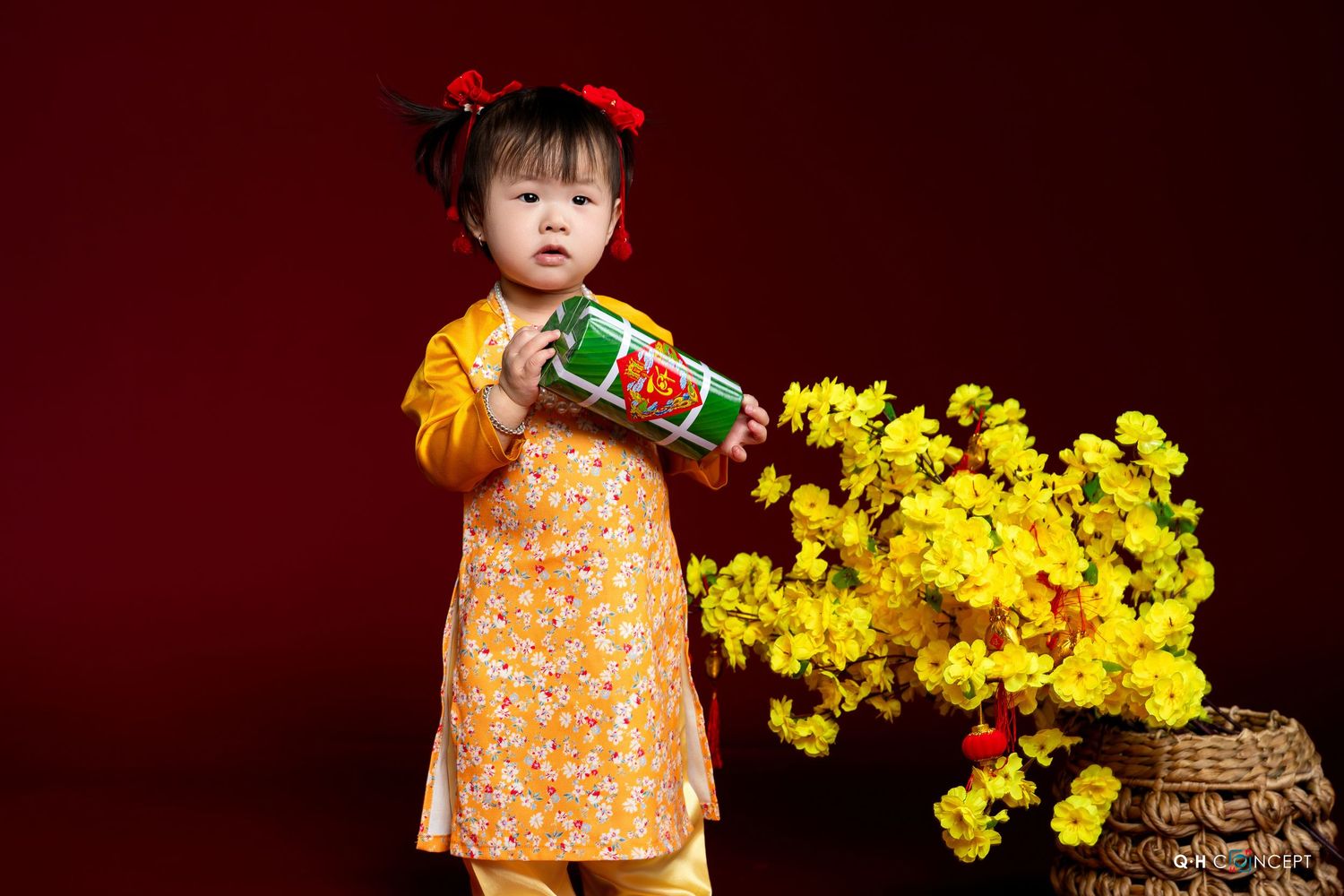 A small child in an orange floral dress arranges bright yellow mimosa flowers in a decorative vase.