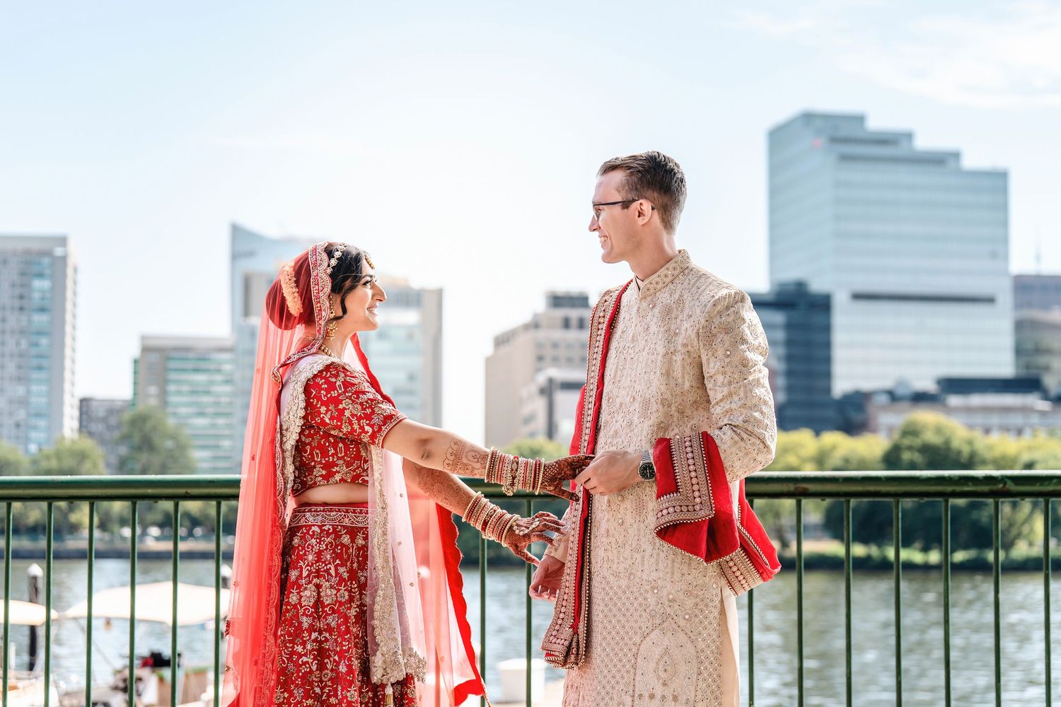 A couple in traditional South Asian wedding attire holds hands on a waterfront terrace with city buildings in the background.