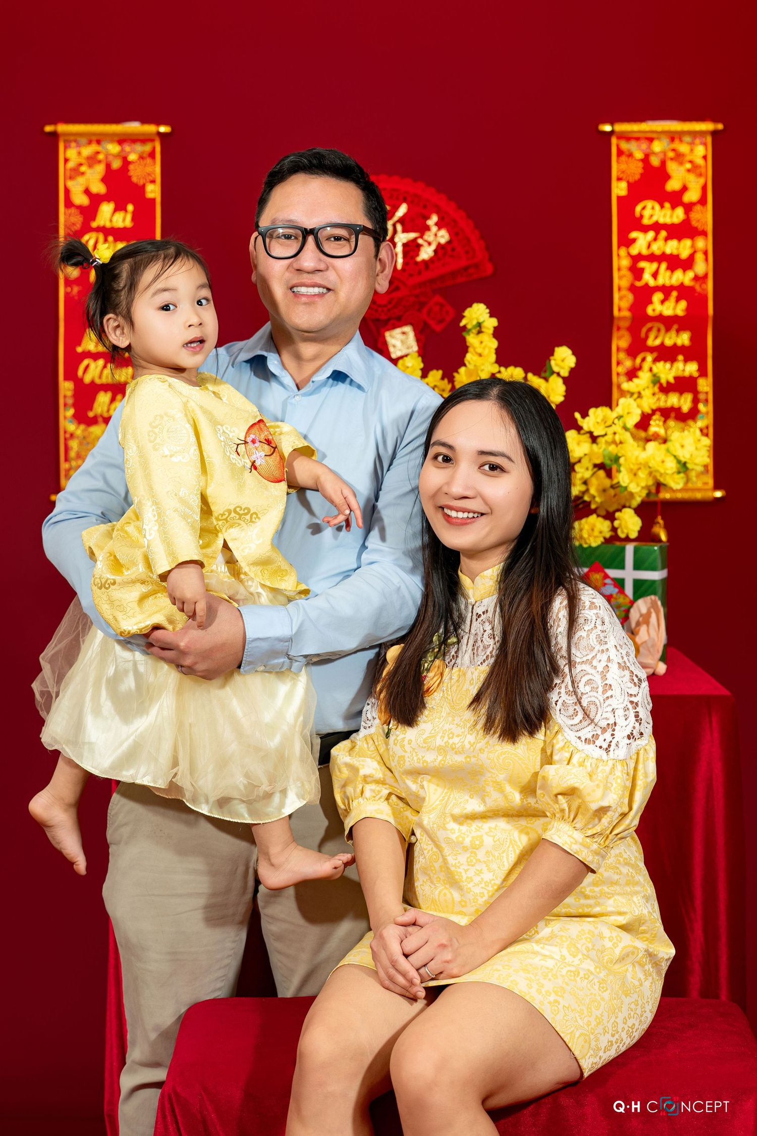 Family poses in traditional Lunar New Year outfits against red backdrop with gold decorations and yellow flowers.
