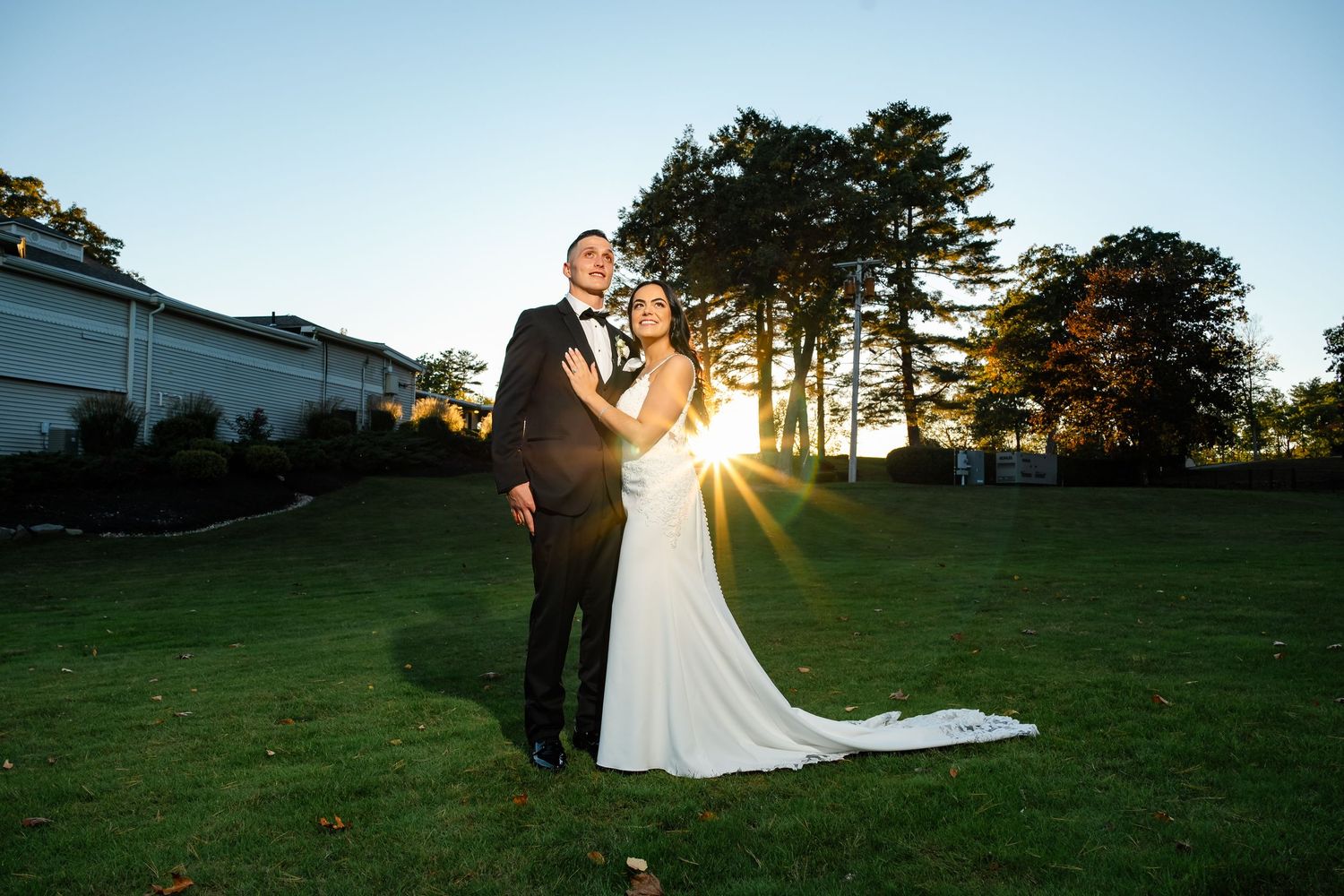 A romantic sunset wedding portrait on a lush green lawn with tall pine trees in the background.