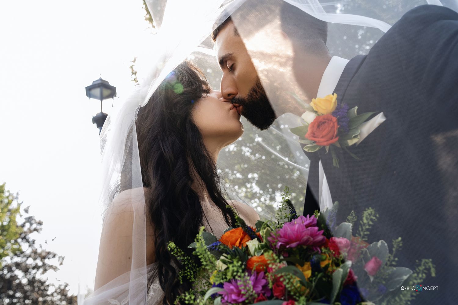 A couple in wedding attire share a romantic kiss while holding a colorful bouquet of flowers outdoors.