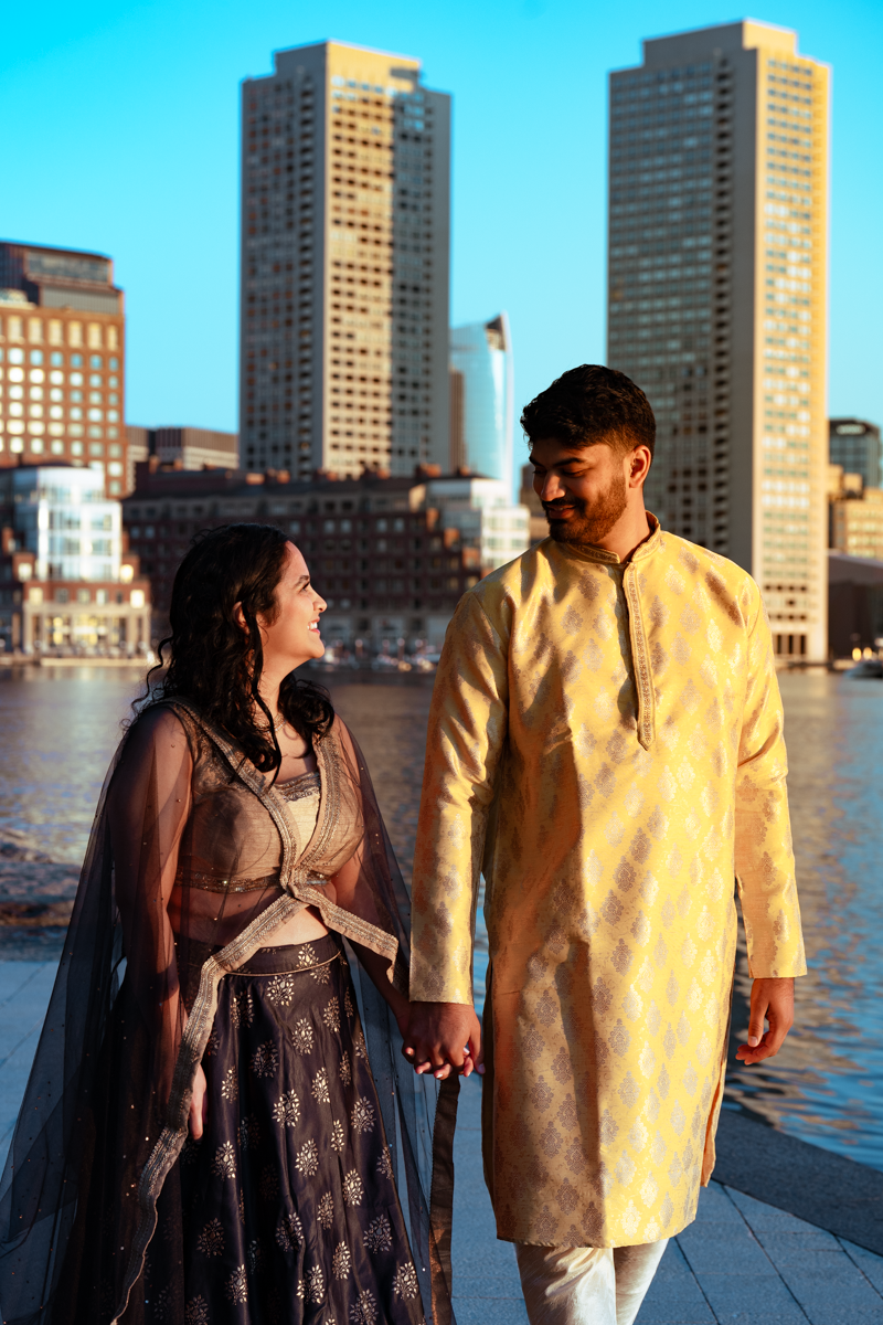 A couple in traditional attire poses by the waterfront with skyscrapers in the background.