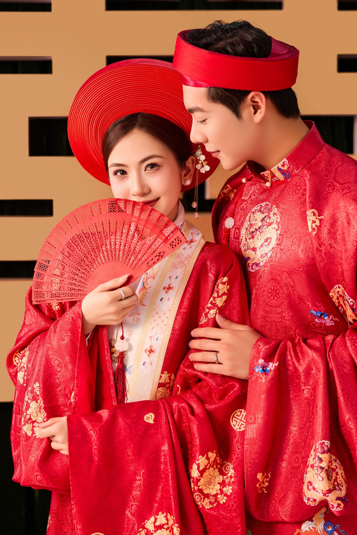 A couple in traditional red Chinese wedding attire share an intimate moment with decorative fans and embroidered details.