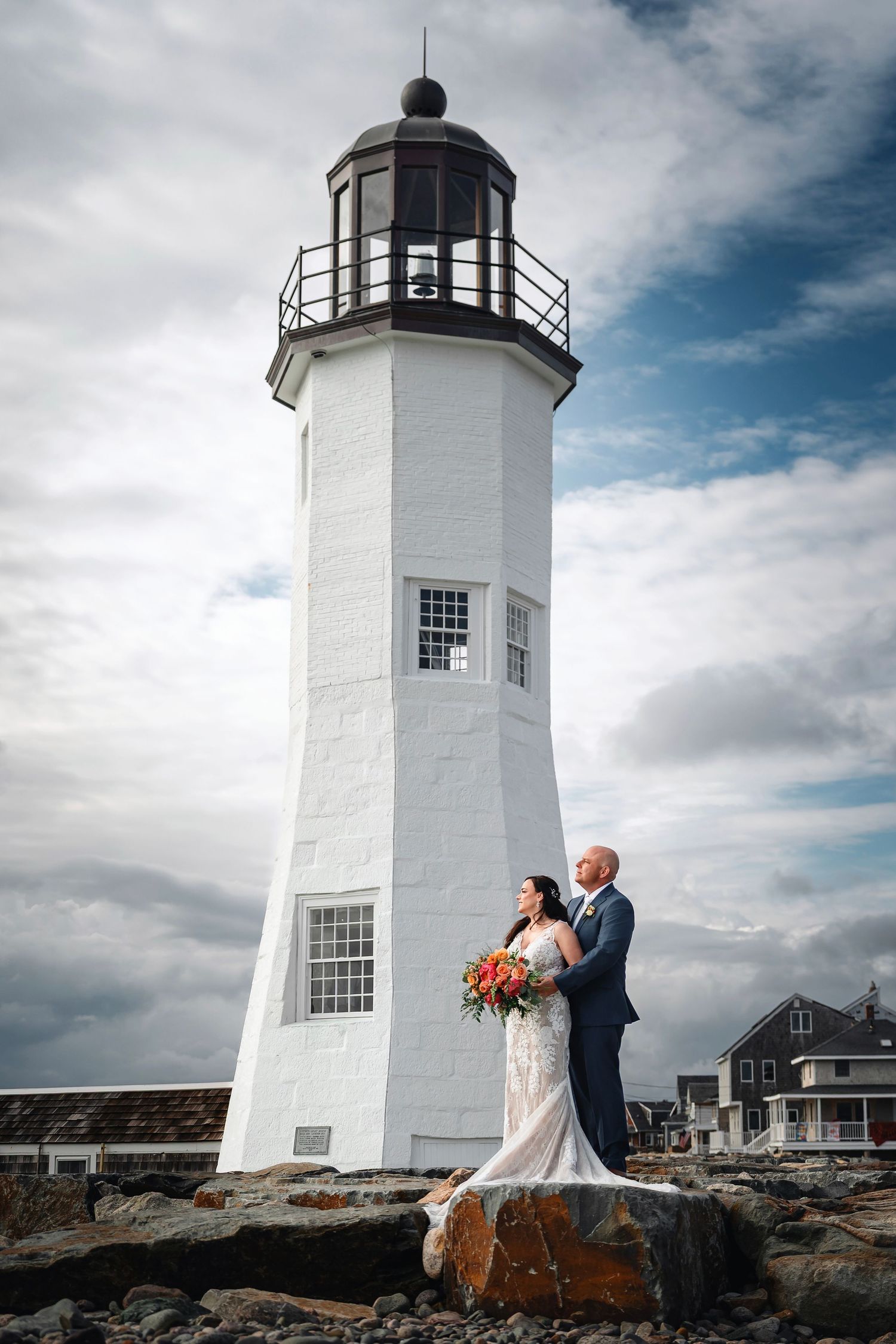 A white lighthouse stands tall against a cloudy sky as a couple poses for a wedding photo on rocky coastal terrain.