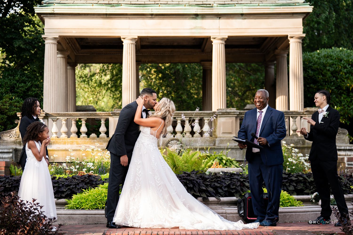 Elegant wedding ceremony taking place under a grand columned pavilion in a garden setting.