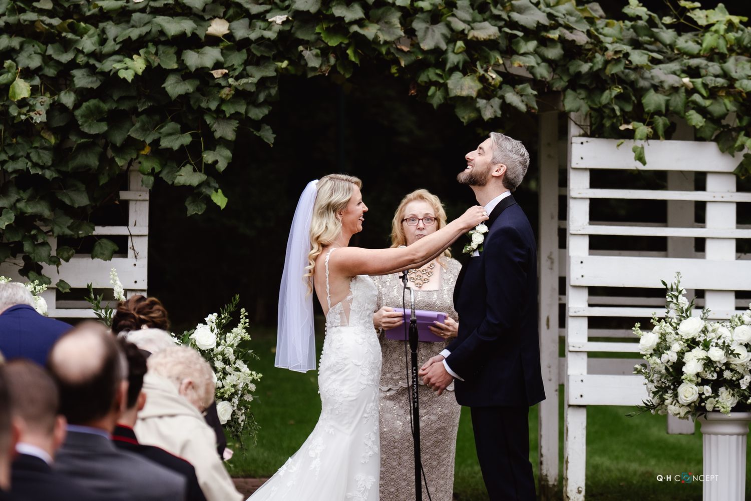A joyful outdoor wedding ceremony takes place in a garden setting with white floral arrangements and ivy-covered walls.