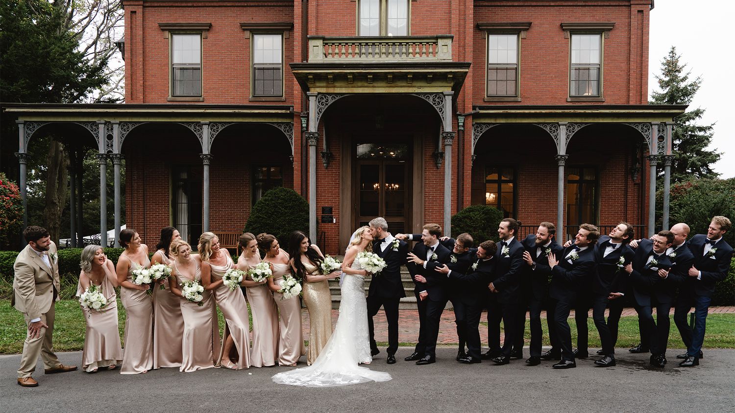 Wedding party poses in a line outside a historic red brick building on a sunny day.