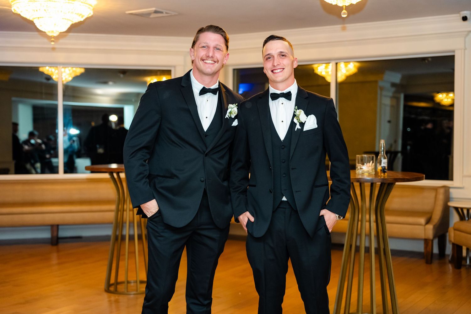 Two people in formal black tuxedos smile in front of mirrored wall.