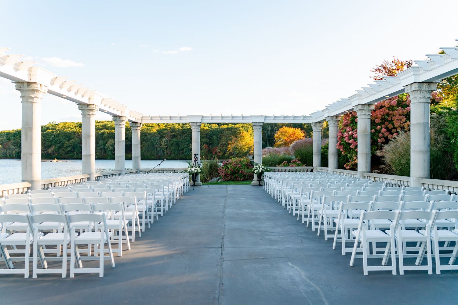 Outdoor wedding ceremony setup with white chairs and pergola overlooking waterfront.