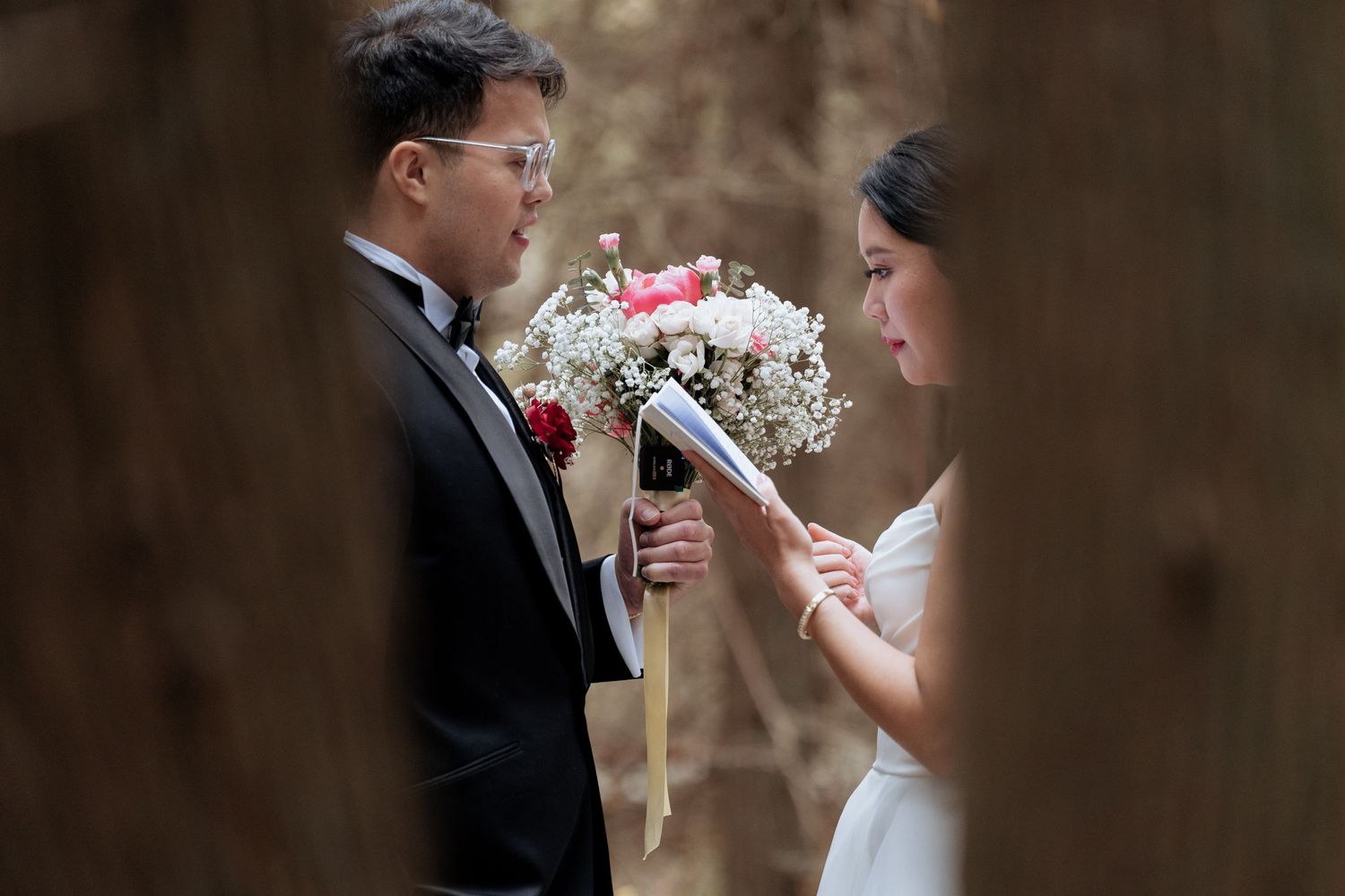 Intimate outdoor wedding portrait captured through trees with soft natural lighting.