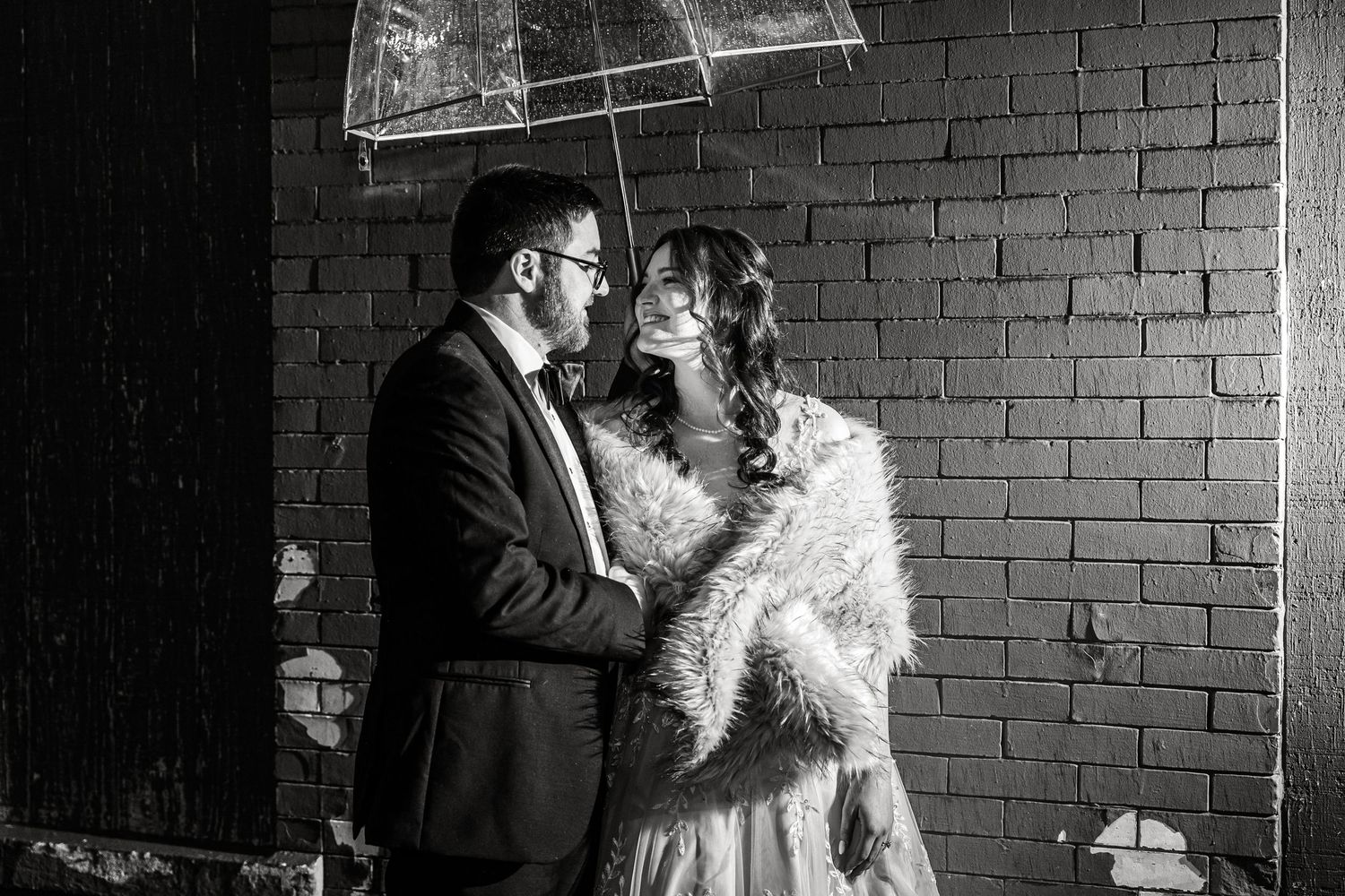Couple sharing an intimate moment under a clear umbrella against brick wall.