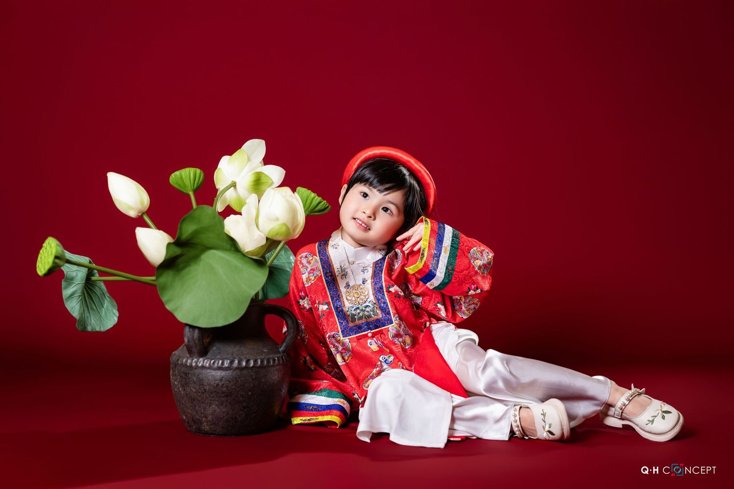 A child wearing a red and white traditional outfit lies down next to white flowers on a red background.