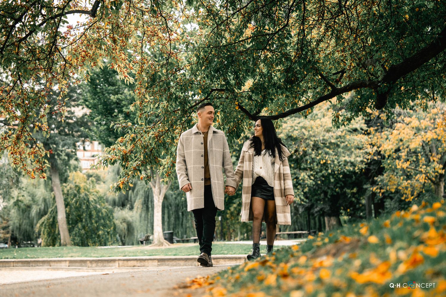 A couple walks through a park pathway lined with blooming yellow flowers and trees.
