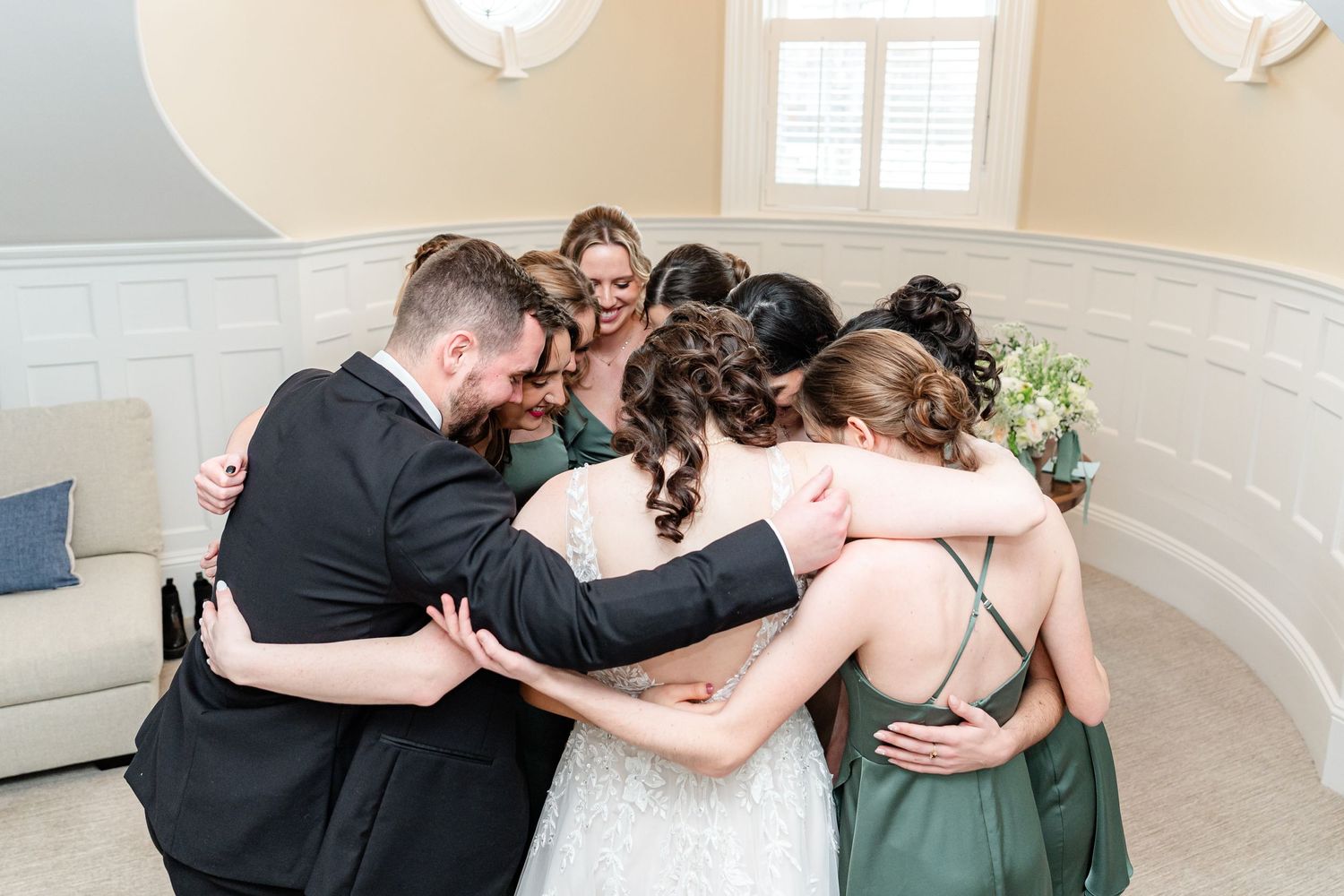 Wedding party embracing in group hug in bright indoor venue.